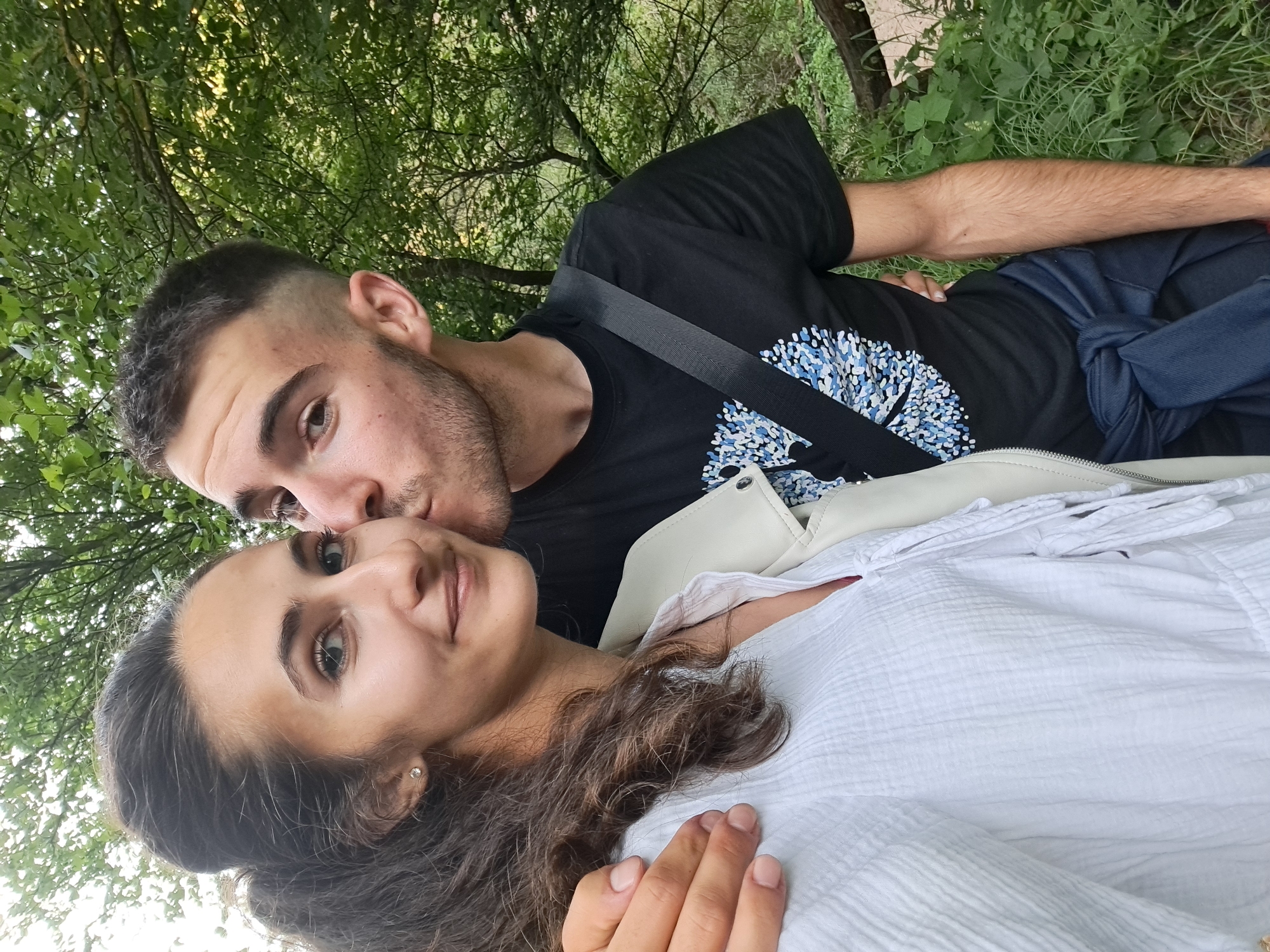 An outdoor, waist-up, selfie-style shot captures a young couple in what appears to be a wooded area in Lokorsko, Bulgaria. The woman, on the left, is facing forward and slightly to the left, with her left arm around the man's waist. She has dark, wavy hair pulled back in a high ponytail, light brown eyes, and is wearing a white textured blouse and a light-colored jacket. A small diamond stud earring is visible in her left ear. The man, on the right, is turned towards the woman and is kissing her on the cheek. He has short, dark hair with an undercut and is wearing a black t-shirt with a blue and white patterned design on the chest and a dark blue jacket tied around his waist. They are positioned in front of lush green foliage and trees, suggesting a natural, possibly rural or park-like setting within Lokorsko. The lighting indicates it is daytime.