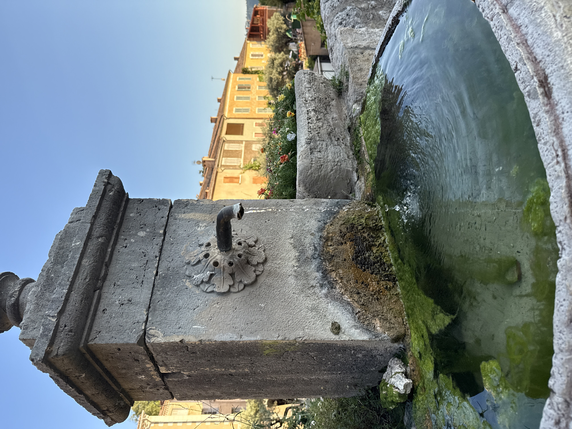 The image shows an old stone fountain with water flowing from a metal spigot into a basin. The fountain is located in Moustiers-Sainte-Marie, France, as evidenced by the background of traditional Provençal buildings. The fountain itself is made of weathered stone, with visible moss and mineral deposits. A decorative floral element surrounds the spigot. In the basin, the water is green with algae, and ripples are visible from the flowing water. Behind the fountain, a stone planter box overflows with colorful flowers, including red, pink, and yellow blossoms. Further in the background, a row of ochre and yellow buildings with tiled roofs lines a street. The sky is a clear blue. The scene appears to be bathed in the warm light of late afternoon or early morning. There are no people visible in the image.