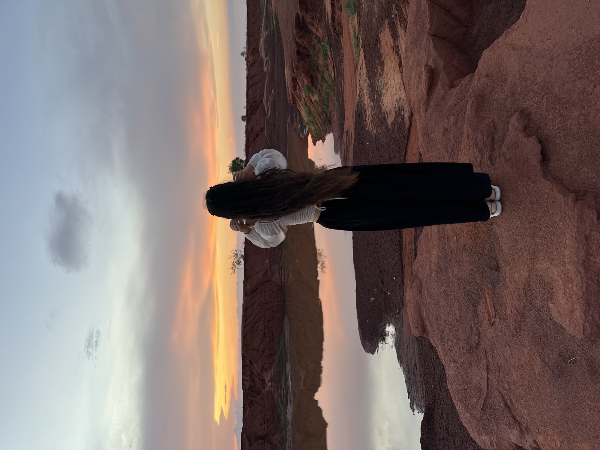 The image depicts a solitary figure admiring a serene natural landscape, most likely during sunrise or sunset, in a location like Mũi Né, Vietnam. The primary subject is a person, viewed from behind, with very long, dark hair extending down their back. They are dressed in a white, long-sleeved top with a subtle texture and a flowing black long skirt or wide-leg trousers, with light-colored shoes visible on their feet.
The person is standing on the edge of a reddish-brown, textured rock formation, which forms the foreground and the right side of the image. Below them and stretching across the middle ground is a calm body of water that acts as a perfect mirror, reflecting the vibrant sky and the surrounding landscape. On the opposite side of this reflective water, more reddish-brown cliffs and hills rise, dotted with sparse green vegetation, hinting at a desert or canyon-like environment.
The sky dominates the upper portion of the image, showcasing a dramatic transition of colors. On the left, the sky is a soft blue-grey with a few scattered clouds, gradually shifting to warm and intense hues of orange, pink, and yellow towards the right, indicative of either dawn or dusk. The soft, warm light bathes the entire scene, highlighting the rich red tones of the earth and creating a tranquil atmosphere. The person appears to be observing the vast, picturesque view. There is no visible text in the image.