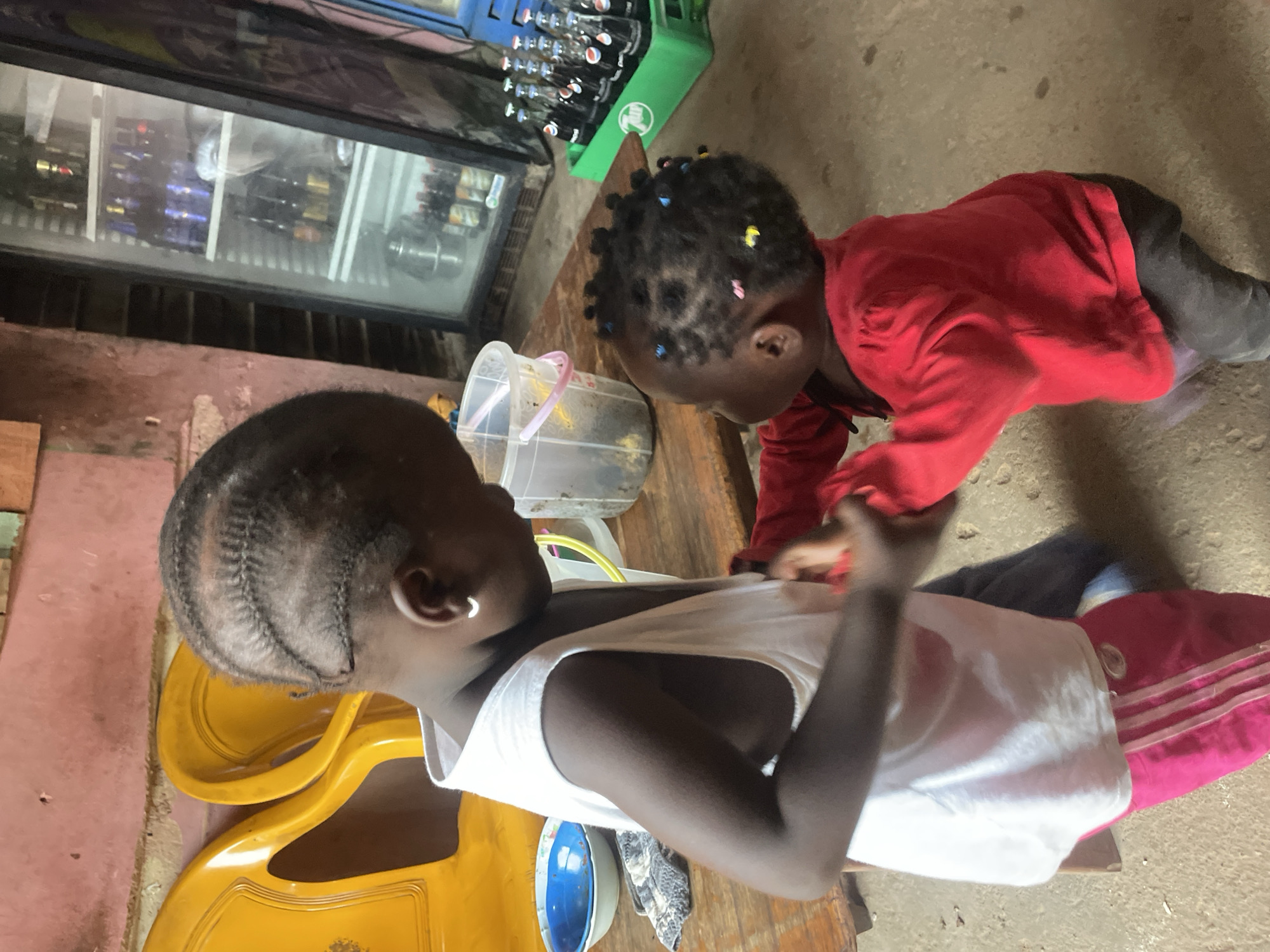 Two young children are interacting at a location in Jos, Nigeria. One child, wearing a white tank top, sits on a yellow plastic chair. The second child, wearing a red long-sleeved shirt, is leaning over a wooden table in front of the first child. On the table, between the children, are a blue bowl, a transparent plastic container with a lid, and other obscured objects. A beverage cooler stands against a wall. A green crate containing bottles of liquid is on top of the cooler. The floor is concrete. A pink and white striped bag is partially visible on the right edge of the frame. The walls appear to be painted in varying shades of red.