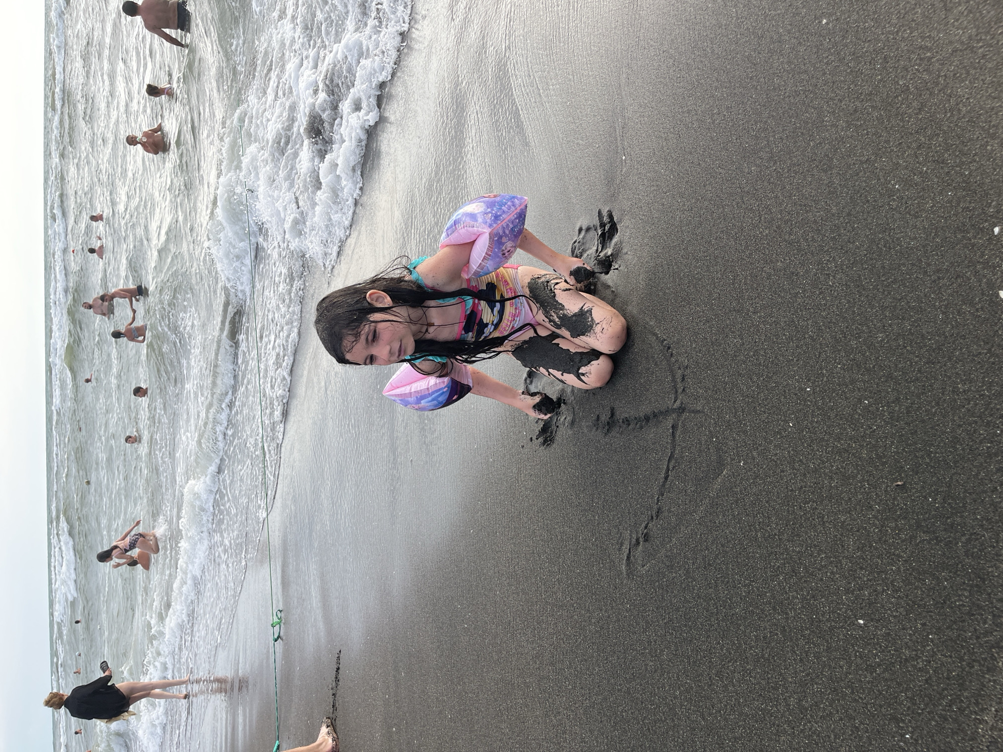 The image shows a child playing with wet sand on a beach. The child is kneeling in the sand, wearing a colorful swimsuit and arm floaties. Their hands are covered in wet sand, and they appear to be molding or digging in it.
In the background, the ocean is visible with gentle waves rolling onto the shore. Several other people are in the water, some swimming and others standing. The overall scene suggests a warm, sunny day at a popular beach destination. The sand appears dark, which is characteristic of some beaches in Georgia.
The focus is on the child's interaction with the sand, conveying a sense of childhood enjoyment and play. The lighting suggests it is daytime, likely afternoon, given the brightness. No visible text is present in the image. The overall emotion conveyed is one of simple, carefree fun in a natural environment.