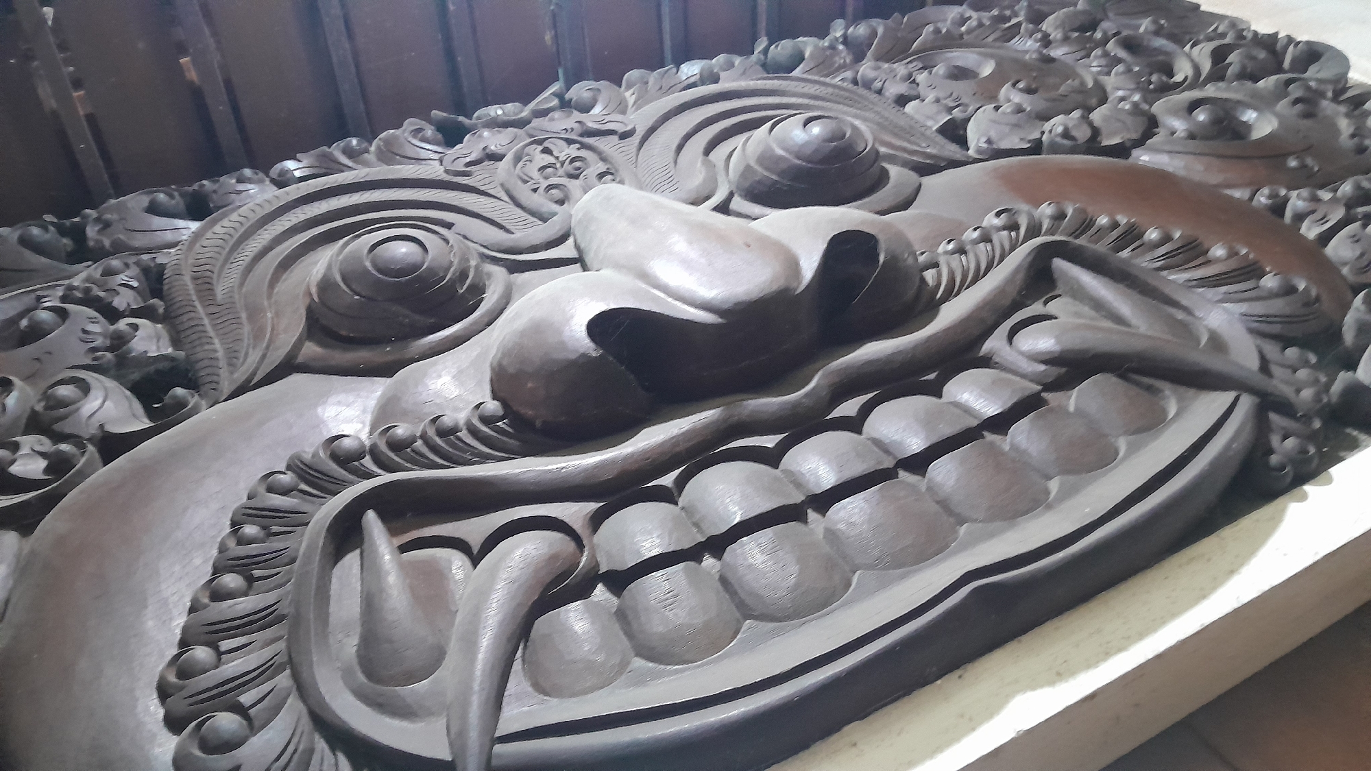The image displays a close-up, eye-level view of a large, intricately carved wooden relief. The carving depicts a monstrous, stylized face, likely a demon or mythological creature, characterized by exaggerated features. Prominent elements include large, bulbous eyes with concentric circles, a wide, gaping mouth filled with numerous rounded teeth, and sharp, pointed fangs. The surrounding ornamentation features swirling patterns, leaf-like motifs, and bead-like details, all rendered in dark, aged wood. The lighting suggests an indoor setting, with shadows emphasizing the depth and texture of the carving. The relief is positioned on a light-colored, flat surface, possibly a pedestal or display area. The background is dark and indistinct, with vertical lines suggesting wooden panels or a wall. No people are visible in the image. The carving is believed to be located in Blora, Indonesia, possibly as a decorative element in a temple, museum, or public space.