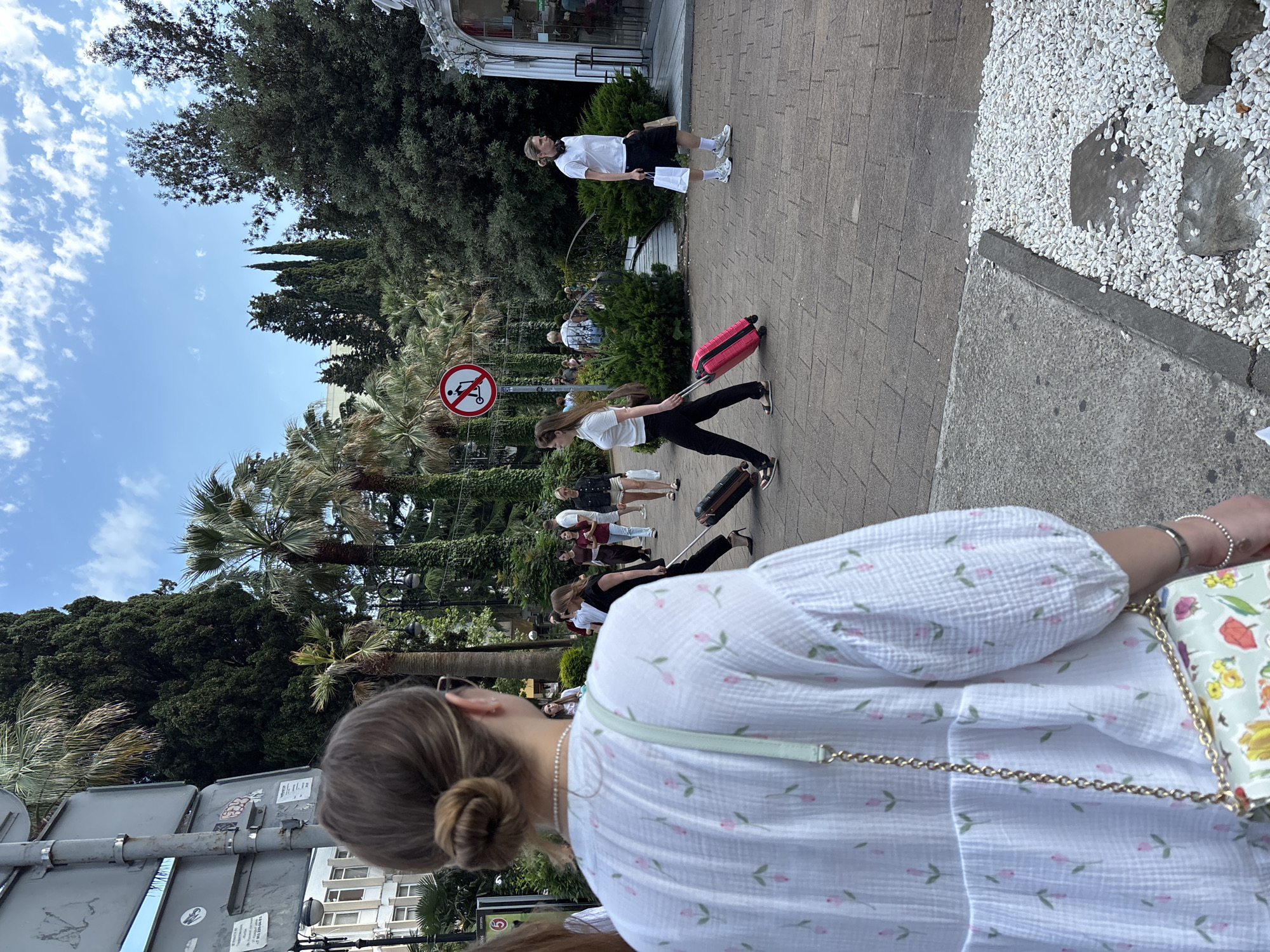 A group of people are walking on a paved walkway lined with palm trees and lush green foliage. A young woman in the foreground is wearing a white patterned dress and is seen from behind, with her hair in a bun. She is carrying a patterned bag with a gold chain. In the middle ground, another young woman is pulling a bright pink suitcase. She is wearing a white t-shirt and black pants. Further back, a person in a white t-shirt and shorts is walking, carrying shopping bags. There are other people in the background, some with luggage. A circular red and white sign with a symbol of a scooter crossed out is visible, indicating no scooters are allowed. The sky is blue with scattered clouds, suggesting it is daytime and the weather is pleasant. The presence of palm trees suggests a warm climate. Buildings are visible in the background. The overall scene appears to be a street or promenade in a resort area. There is some indecipherable text on stickers on a metal pole in the foreground.