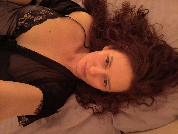 A single individual, an adult female with long, dark reddish-brown curly hair, is depicted lying down indoors. She is positioned diagonally across the frame, looking directly at the camera with a neutral expression. Her hair is spread out on a light-colored, soft surface, likely a pillow on a bed. She is wearing a black, sheer garment, appearing to be a robe or negligee, which features lace detailing over her left breast and on the trim near her left arm. A small, dark mole is visible on her upper torso, situated above her left breast. The scene is illuminated by warm, soft lighting, casting an orange-pink hue over her skin and the surrounding light-colored bedding. The background is indistinct. The precise location within the city of Saint Petersburg, Russia, cannot be determined from the visual evidence provided in the image.
