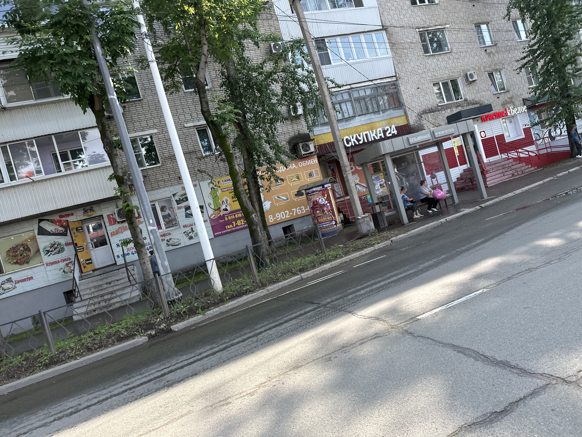 A street view in Birobidzhan, Russia, shows a light-colored brick building with several shops on the ground floor, partially obscured by trees. The shops have various signage, including what appears to be a food establishment with images of shawarma and other dishes, and another with text that may read "СКУПКА 24" (meaning "Buyout 24"). A street runs along the left side of the image, with cracked asphalt and faint tire tracks. Several people are visible near the entrance of one of the shops, with one person sitting on a bench and another standing. The overall scene suggests a commercial area within the city.