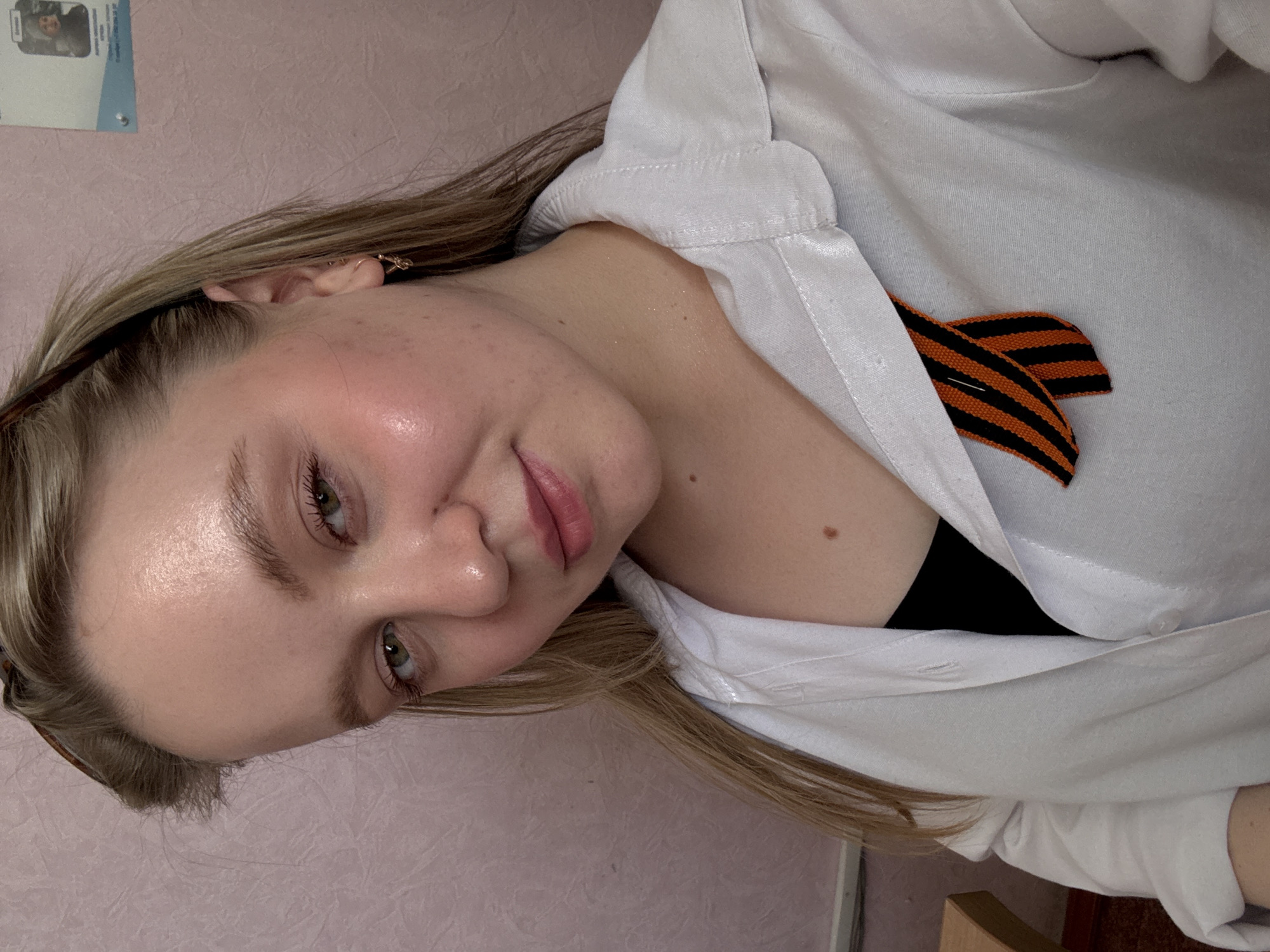 A close-up shot of a young woman. She has fair skin, blonde hair, and green eyes. She is wearing a white button-down shirt. Pinned to her shirt is a St. George ribbon. A small mole is visible on her neck. The background is a light pink wall. Part of a document is visible in the top left corner. The scene appears to be indoors. The city is Chaikovsky, Russia.