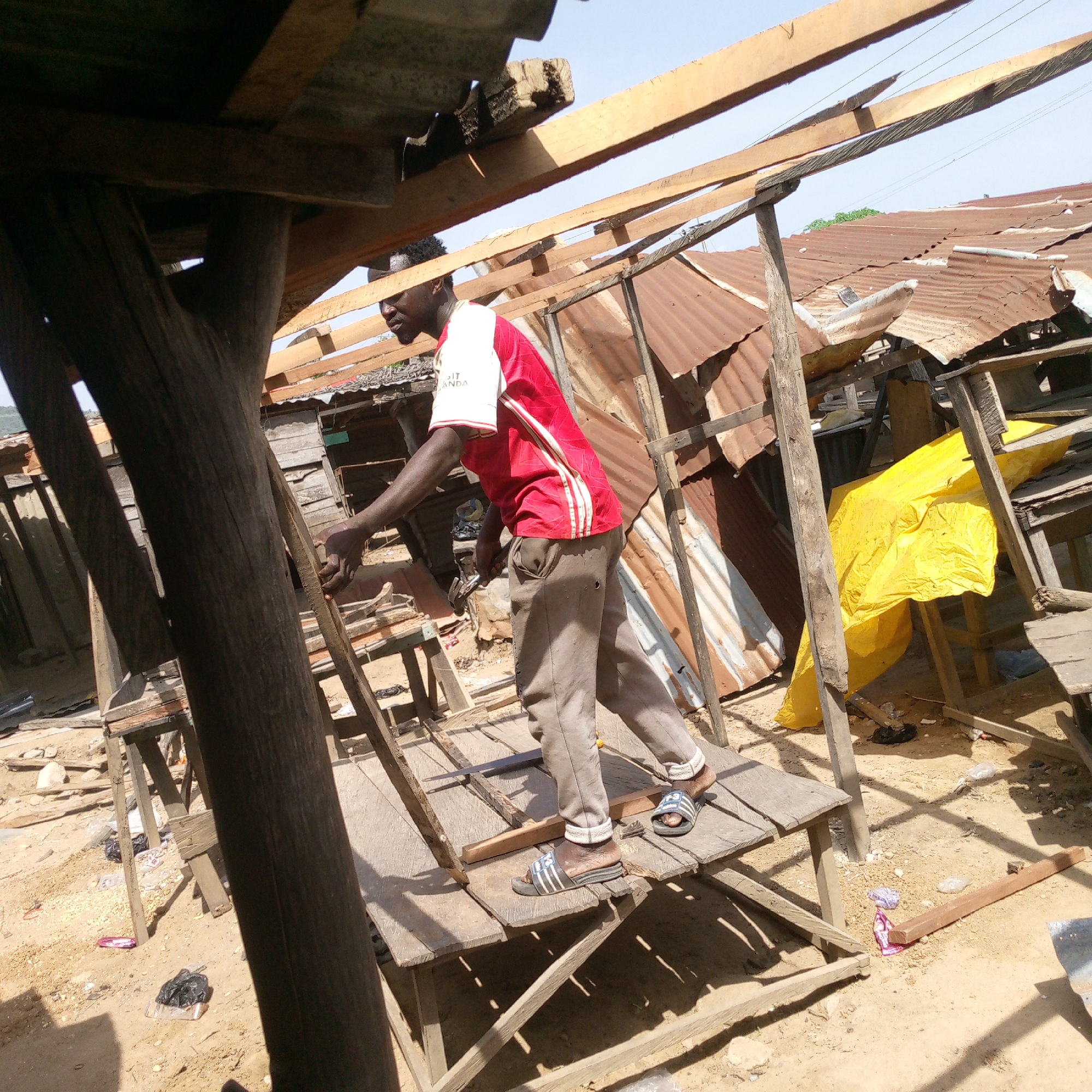 This image, captured in Adankolo, Nigeria, depicts a man engaged in construction or repair work under bright daylight. The subject is a Black man, seen from the side, standing on a basic wooden platform. He wears a red and white short-sleeved shirt with the partial text "SIT ANDA" visible, grey trousers, and black and white striped slide sandals. In his right hand, he holds a hammer, appearing to work on a wooden beam or plank.
The scene is an outdoor setting characterized by rudimentary wooden structures, likely stalls or sheds. These structures feature exposed wooden posts and beams. In the background, numerous older buildings with rusty and damaged corrugated metal roofs are visible, interspersed with newer wooden rafters, suggesting ongoing construction or renovation. A bright yellow tarp is gathered beneath one of the structures. The ground is sandy, typical of a dirt path or undeveloped area, and shows scattered debris such as plastic bags and wrappers. Strong shadows cast by the structures and the man indicate a sunny day, likely midday or early afternoon. The overall atmosphere suggests an active, informal construction environment, possibly within a market or developing area.