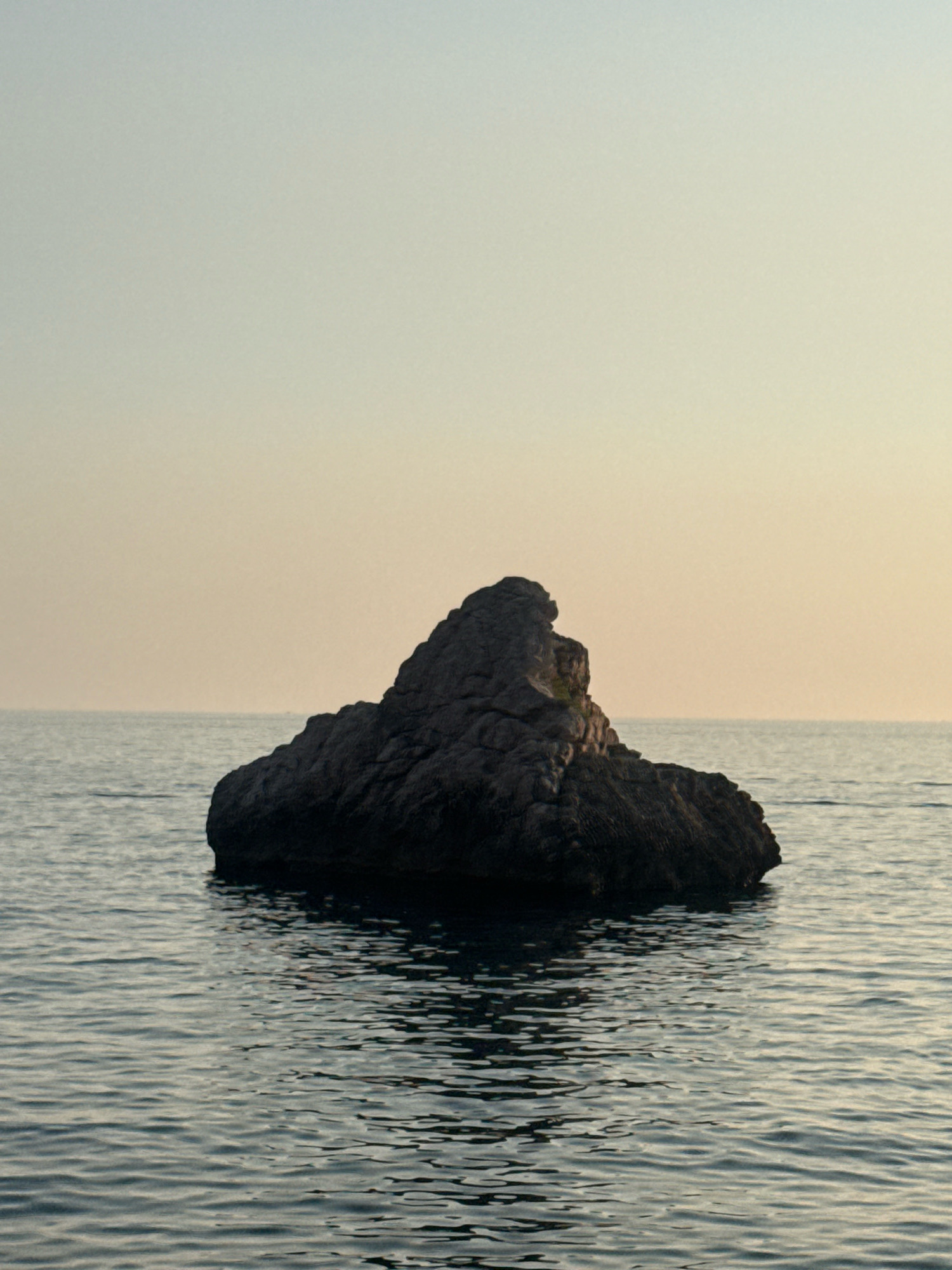 A solitary, dark, irregularly shaped rock formation emerges from the calm, rippling surface of the Adriatic Sea. The rock, which appears to be a significant size, is dark gray or black and has a rugged, textured surface. It is positioned centrally in the frame, with the sea extending to the horizon. The water is a deep blue-gray, reflecting the muted tones of the sky. The sky itself is a gradient of pale blue at the top, transitioning to a warm, hazy yellow or light orange near the horizon, suggesting either sunrise or sunset. The scene is tranquil and devoid of any human presence or activity. The rock formation is likely a natural landmark, possibly part of the coastline or an offshore reef near Rafailovići, Montenegro, given the characteristic clear waters and coastal rock formations of the region.