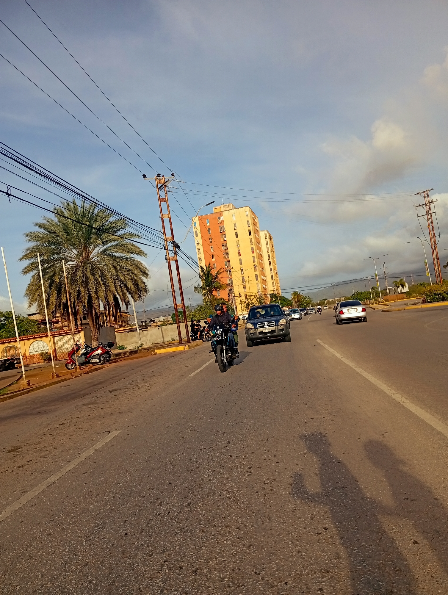 The image depicts a street scene in Juan Griego, Venezuela, under a partly cloudy sky. The perspective is from a moving vehicle, capturing a road with multiple lanes. In the foreground, the asphalt road shows some wear and tear, with a white line marking a lane division. Two distinct shadows of people are cast on the road surface, indicating the presence of the photographer and potentially another individual.
On the left side of the road, there are parked motorcycles and a palm tree. Further down the street, a dark-colored SUV is in motion, followed by other vehicles, including a white sedan. In the background, several tall, light-colored apartment buildings are visible, characteristic of urban architecture. Utility poles with numerous electrical wires stretch across the sky, a common sight in many cities. The overall impression is of a typical urban environment during daylight hours, with moderate traffic and infrastructure.