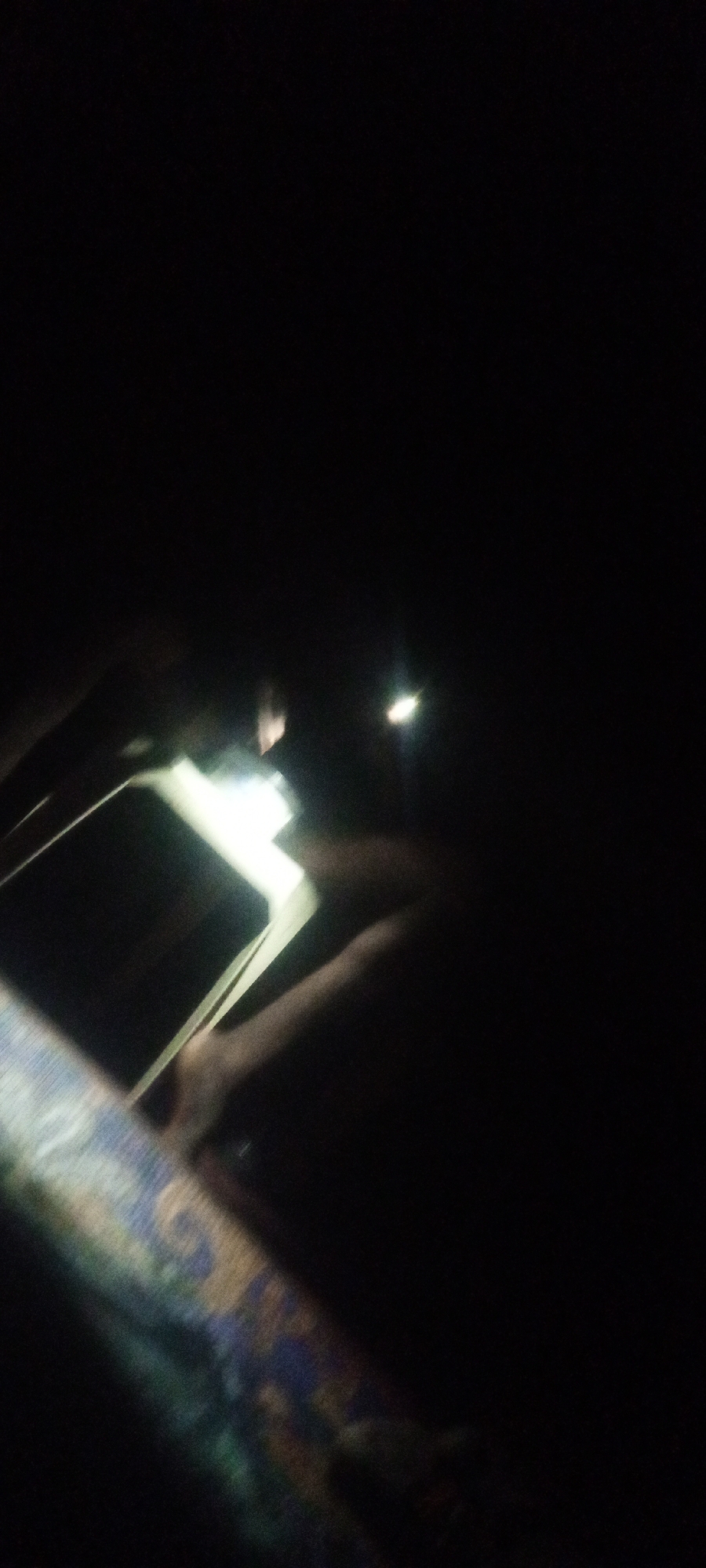 In a dark environment, a person is holding a book, illuminating it with a light source from a phone or flashlight. The book appears to be lying on a patterned surface, possibly a blanket or piece of furniture. The scene is dimly lit, with most details obscured by darkness. Location is unknown.