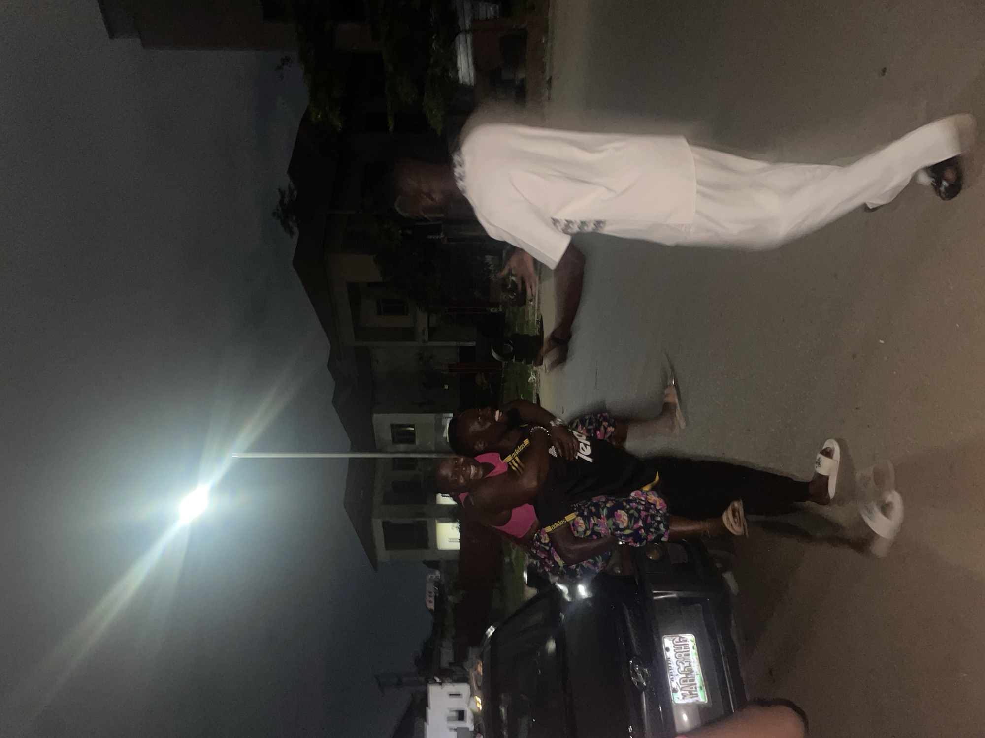 In Guzape, Nigeria, at night, a man gives a woman a piggyback ride. The woman, wearing a pink top and floral-patterned pants, laughs while on the man's back, who is wearing a black shirt and similar pants. Another man in a white outfit stands in front of them, with his arms extended and hands open, looking towards them. A dark-colored car is parked to the left of the frame, and buildings are visible in the background under the glare of a street lamp.