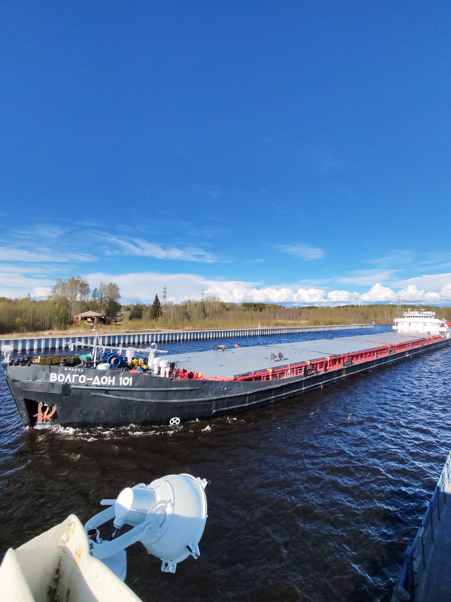 A barge named "VOLGO-DON 101" is moving through a canal. The barge is black with red accents and a gray deck. There are trees and some buildings on the canal bank. The sky is blue with white clouds. The image appears to have been taken from another vessel. The location is Анхимово, Russia.