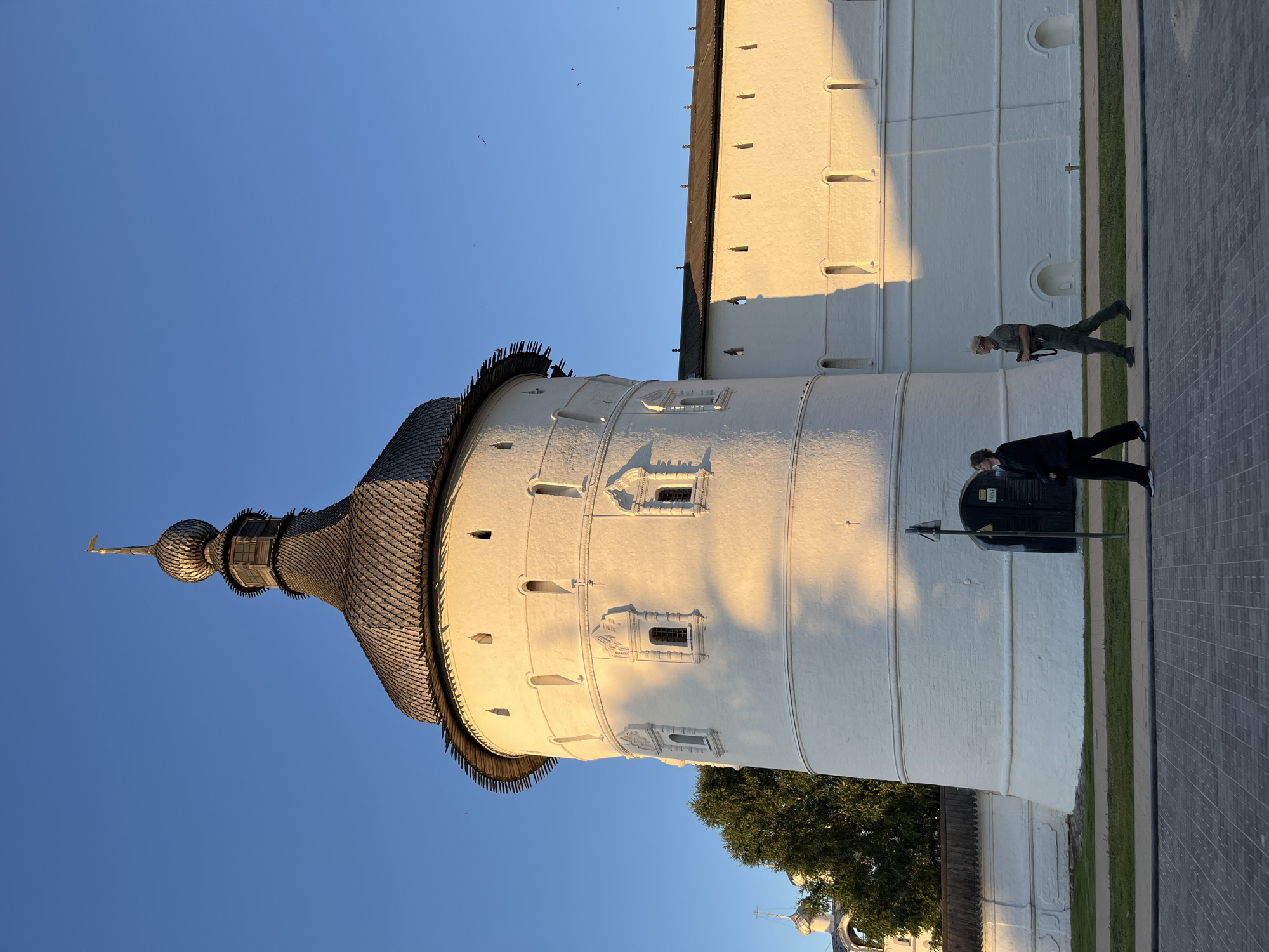 The image depicts a section of a white, cylindrical tower with a dark, conical roof, likely part of a religious or historical complex. The tower has numerous small, rectangular windows and decorative elements. Sunlight casts long shadows across the textured white surface, indicating it is either early morning or late afternoon. The sky is a clear, bright blue, suggesting fair weather.
Two individuals are visible in the scene, walking on a paved area adjacent to the tower. One person, wearing a light-colored hat and olive-green clothing, is closer to the tower. Another person in a dark suit and tie is walking further along the path. Their presence suggests the location is open to visitors.
A dense green tree is partially visible behind the tower, its foliage contrasting with the stark white of the building. In the distance, a glimpse of another building with a dome and cross can be seen, reinforcing the impression of a religious site. The paving stones are arranged in a patterned grid.
The image is oriented vertically, with the tower appearing to lean to the left. There is no discernible text within the image. The overall impression is one of a historical landmark bathed in warm, golden light, with a sense of calm and timelessness.