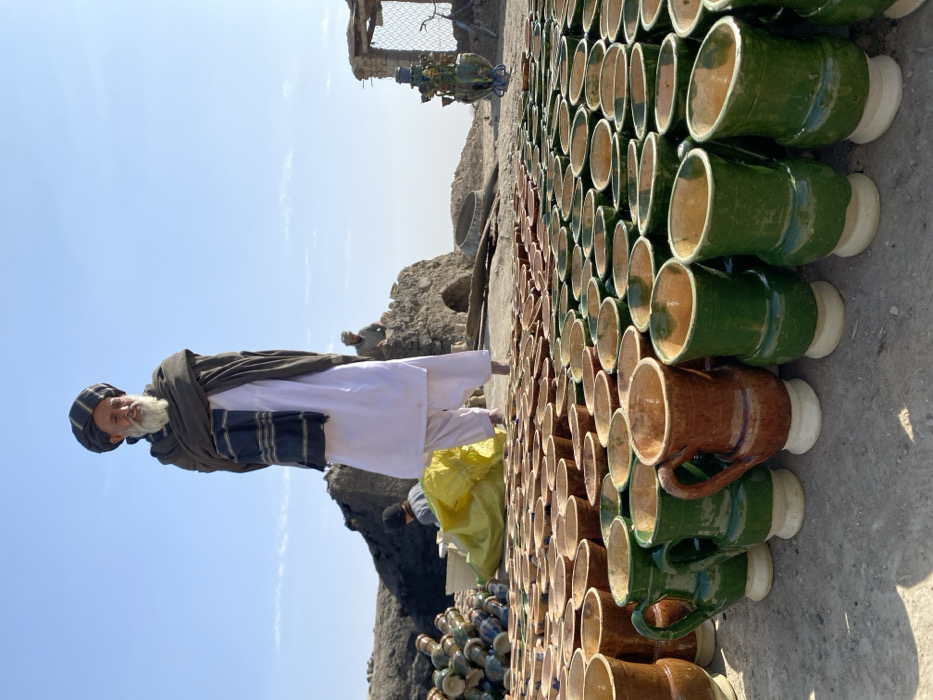 The image portrays an outdoor pottery production area in Kandahar, Afghanistan, under a clear, bright blue sky. In the foreground, hundreds of newly crafted ceramic mugs and cups are meticulously arranged in long rows on the bare, dusty ground, drying in the open air. These vessels display a predominant color scheme of earthy brown and dark green glazes, with unglazed, light-colored bases.
A prominent elderly man stands in the mid-left of the frame, facing slightly right. He has a long, white beard and is dressed in traditional attire: a dark, wrapped turban, a dark shawl with striped patterns draped over his shoulders, a loose-fitting white tunic (qameez), and white trousers (shalwar). His hands are tucked into his shawl.
In the mid-ground and background, the scene reveals further aspects of the pottery operation. To the far left, a stack of glazed pottery pieces, exhibiting blue and green tones, is visible. Further back, two other men are present near rugged, possibly mud-brick or stone structures, which appear to be kilns or workshops. One man is partially obscured behind a bright yellow tarp or bag, while another sits atop a low, weathered structure to the right of the main figure. To the far right, a large, ornate, green-glazed ceramic vessel stands upright near a dilapidated structure that includes some wire fencing. The overall environment is arid and dusty, indicative of an outdoor artisan's workspace.