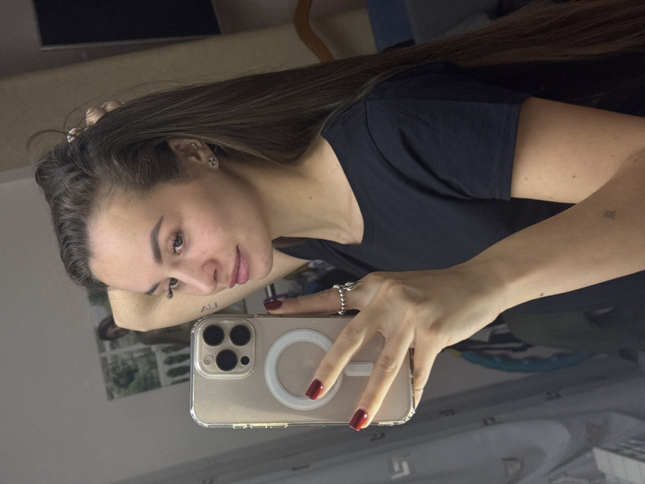 A woman with long brown hair poses for a selfie in a mirror. She wears a dark t-shirt and has red nail polish. Her phone case is clear with a white ring on the back. A small star tattoo is visible on her arm. She wears stud earrings. The background reflects a room interior and window with greenery outside.