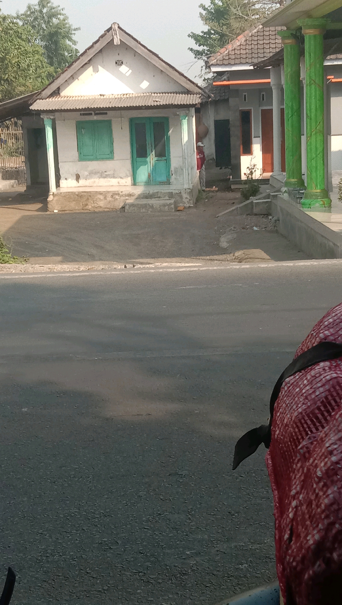 The image depicts a street scene in Karang Anyar, Indonesia, with a focus on two adjacent buildings. The building on the left is a small, single-story structure with a white facade and a gabled roof covered in corrugated metal. It features two teal-colored doors and one teal window. The building on the right is larger and appears to be a shop or commercial establishment with a tiled roof and prominent green columns at its entrance. The street in the foreground is asphalt, and a portion of a person's red and white checkered garment and a black strap are visible in the lower right corner, suggesting the photo was taken from a moving vehicle or from behind someone. The environment appears to be a sunny day with some shadows cast on the road.