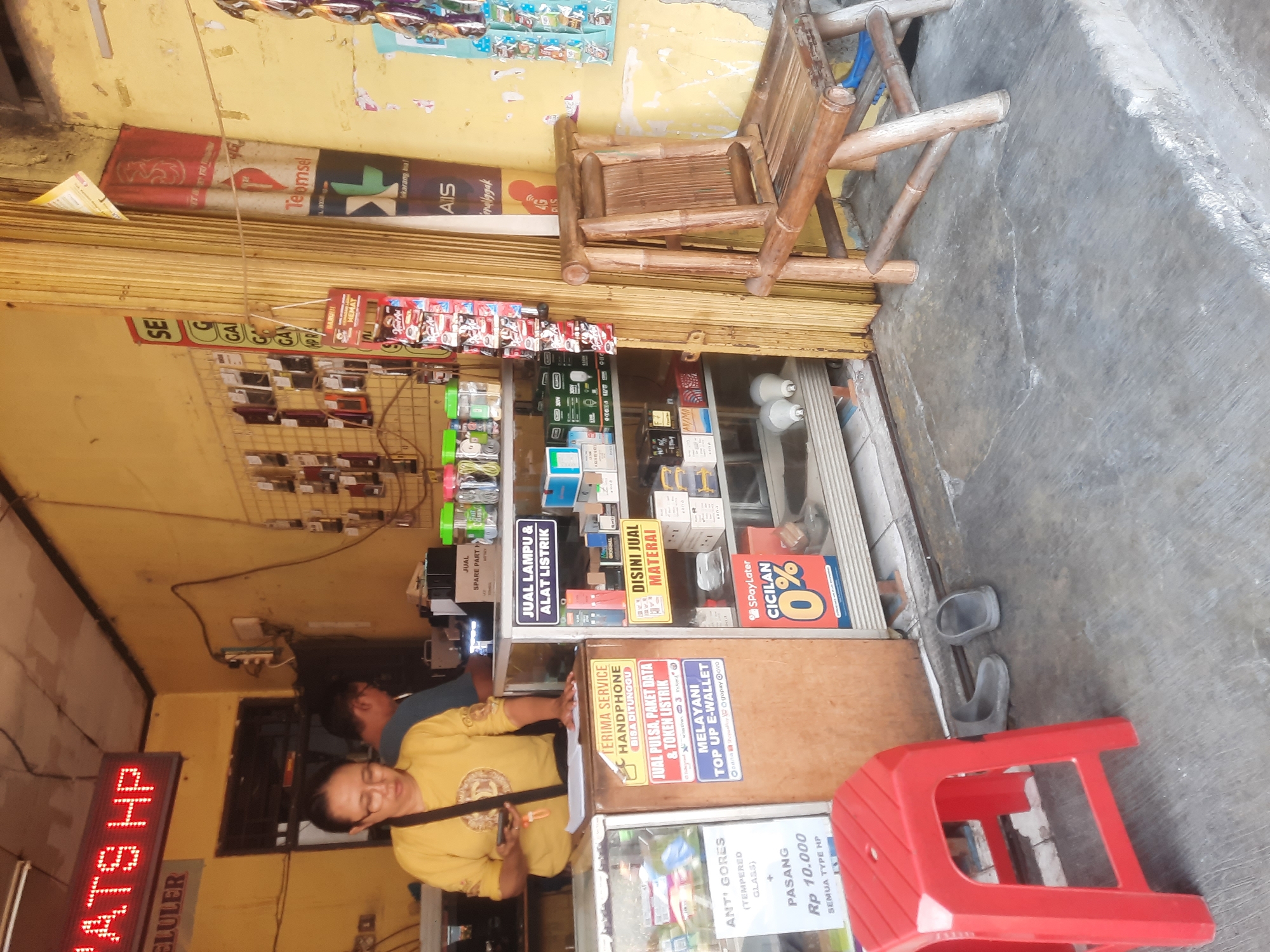 A medium shot captures an outdoor street-level storefront in North Jakarta, Indonesia. The storefront appears to be a small, informal shop selling mobile phone accessories and services. The facade is painted a vibrant yellow, with some peeling paint visible. A wooden door is folded open to the right.
In the foreground, a red plastic stool is placed on a gray concrete floor, with a pair of dark gray sandals next to it.
Inside the shop, a woman wearing glasses and a yellow long-sleeved shirt stands behind a glass counter, holding a smartphone and looking down at it. She appears to be interacting with the phone or a transaction. To her left, a man in a blue shirt is partially visible, seemingly working behind the counter.
The counter is filled with various items, including shelves stocked with small boxes, packaging, and a display of mobile phone accessories. Several signs are attached to the counter and the wall. One sign in English and Indonesian advertises "ANTI-GORES (TEMPERED GLASS)" with a price of "Rp 10.000". Another sign in Indonesian reads "TERIMA SERVICE HANDPHONE BISA DITUNGGU" (We accept phone service, can be waited for). A sign above a shelf in the shop reads "JUAL LAMPU & ALAT LISTRIK" (Selling lamps and electrical equipment). Another sign on the wall promotes "SPayLater CICILAN 0%" (SPayLater Installment 0%).
Prominently displayed on a rack to the right of the counter are various small packages of goods, possibly snacks or small electronic accessories. To the far right, a bamboo chair with a woven seat and back is positioned near the folded wooden door.
Above the counter, a digital sign displays "ATS HP" in red LED lights. To the left of this, a sign reads "ELULER". Further to the right, larger signs are visible, including one with the "XL" logo and another with the "Telkomsel" logo, indicating the presence of telecommunication services. The overall scene suggests a busy, everyday retail environment in a bustling Indonesian city.