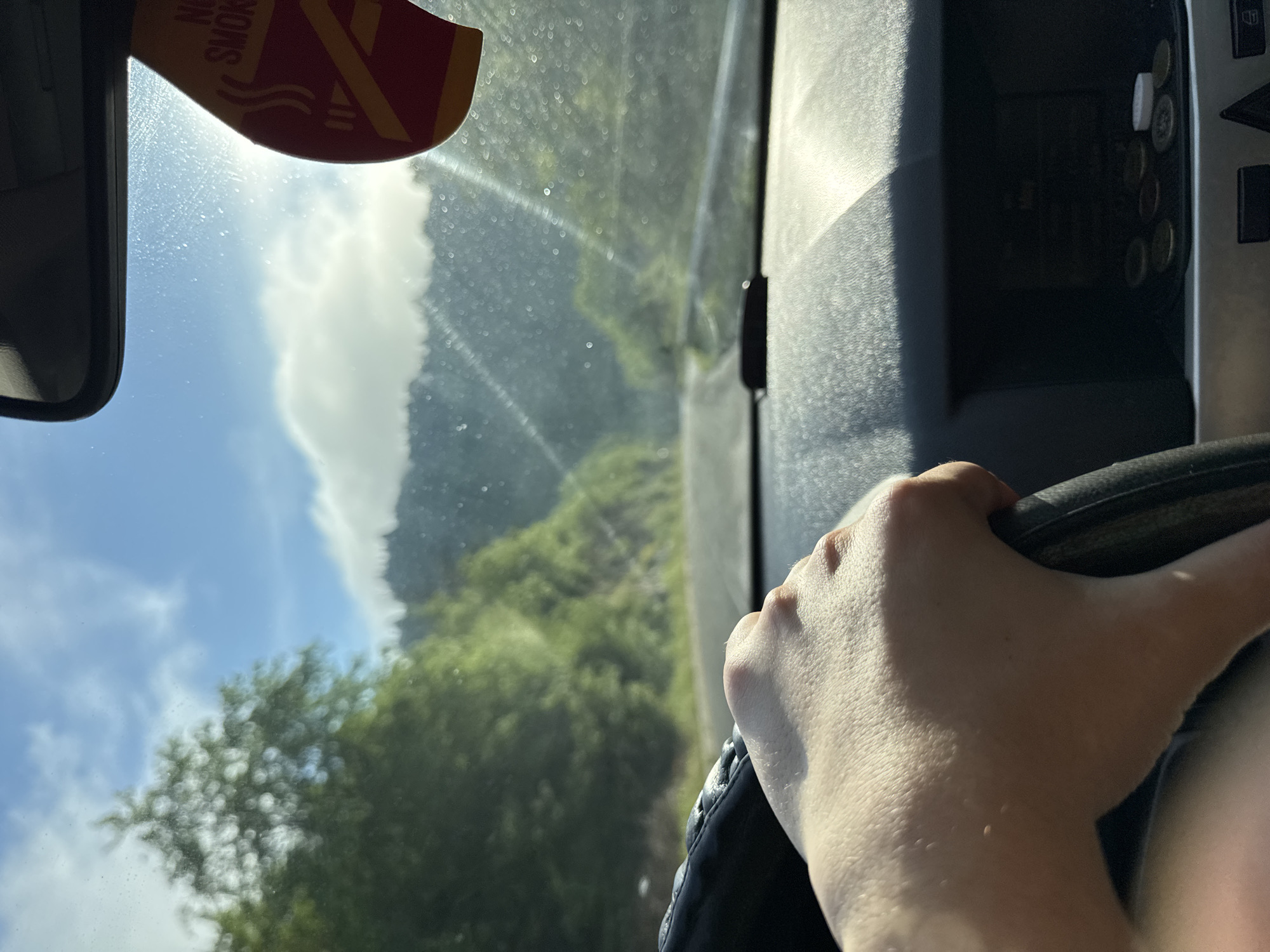 The image shows a view from inside a vehicle, presumably a car, with a hand on the steering wheel. The windshield offers a blurred view of a winding road lined with dense green trees and vegetation, suggesting a mountainous or hilly area. In the distance, there are hints of a cloudy sky with bright sunlight. A "No Smoking" sticker is adhered to the upper left portion of the windshield. To the right of the windshield, there are some console buttons and possibly coins in a slot. The location in the city of Taran, Bulgaria is not discernable from the visual information provided.