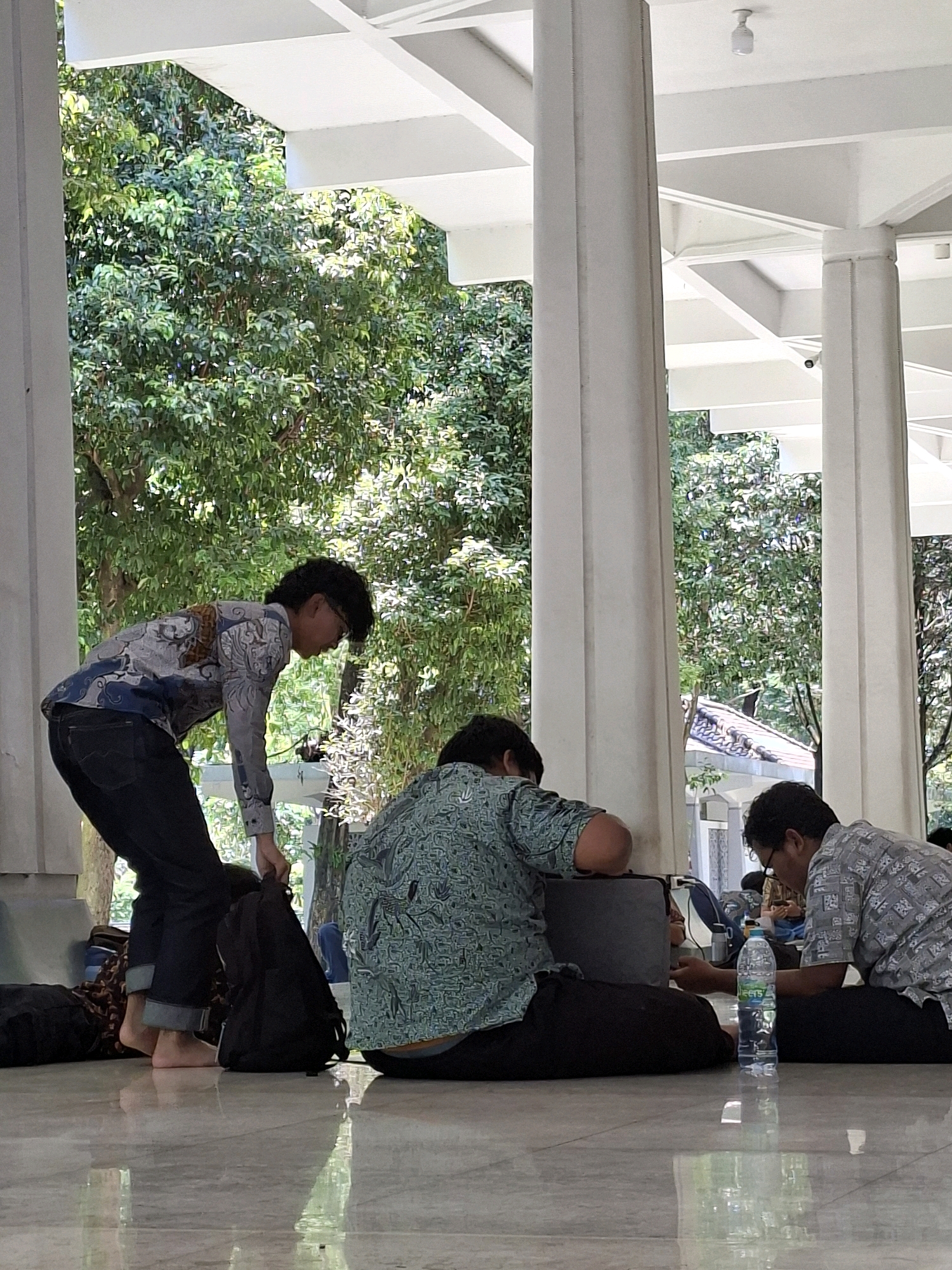 In an open-air covered area with white pillars and a white ceiling, several people are sitting and standing on a polished, reflective floor. In the foreground, a person wearing a patterned blue and white long-sleeved shirt and dark jeans is bending over, holding a black backpack. To their right, a person in a green patterned long-sleeved shirt and black pants is leaning forward, resting their head on a dark object, possibly a laptop. Further to the right, another person in a grey patterned long-sleeved shirt and black pants is looking down at something on the floor. A bottle of water is placed on the floor in front of them. Lush green trees are visible in the background, suggesting an outdoor or semi-outdoor location. The setting appears to be a public space, possibly within a university campus or a recreational area. The overall atmosphere suggests a moment of rest or study. This scene is located in the city of Surabaya, Indonesia.