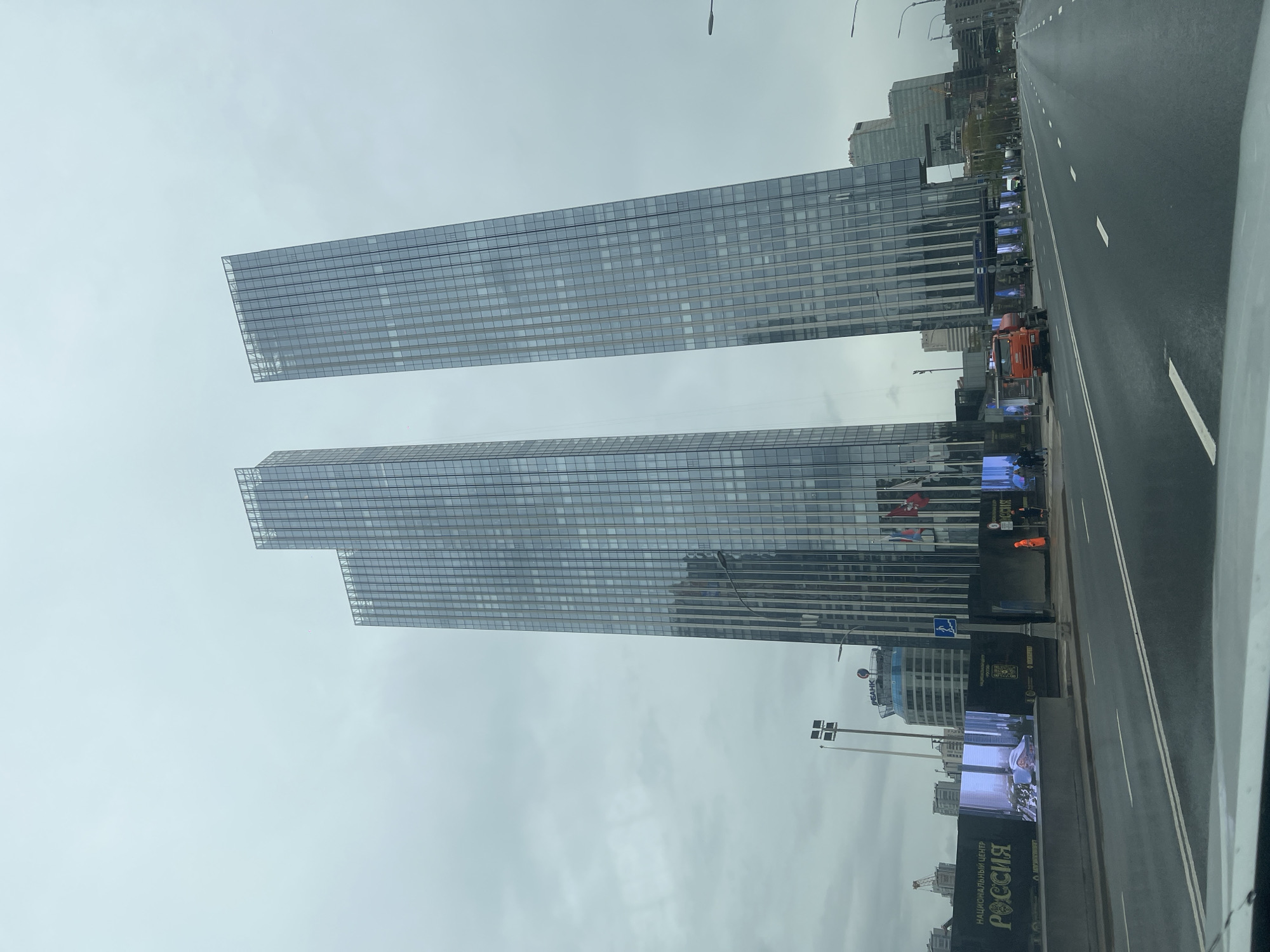 The image shows two modern glass-clad skyscrapers in Moscow, Russia, likely near the Moscow International Business Center (Moscow City). The buildings are rectangular in shape and feature a grid-like pattern of windows. A road with white lane markings runs along the right side of the image. Digital billboards are visible along the roadside. The sky is overcast. The angle suggests the image was taken from a vehicle.