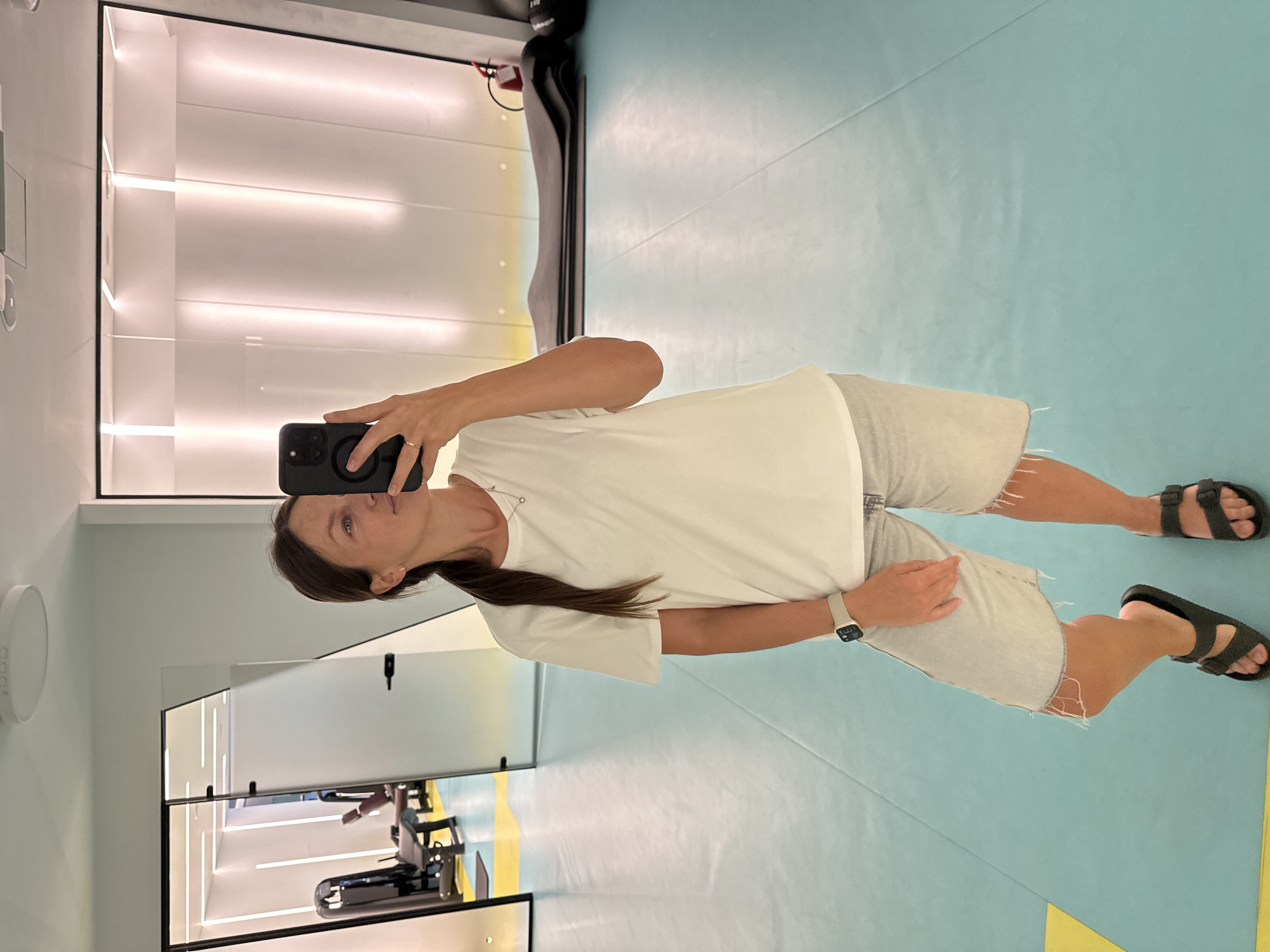 A woman is taking a mirror selfie in a gym located in Boreti, Montenegro. She is wearing a white t-shirt, light-wash denim shorts with distressed hems, and black sandals. She has a watch on her left wrist and a necklace with a small pendant. Her right hand is holding a smartphone. The gym has a light blue floor with yellow markings. In the background, through a glass door, fitness equipment is visible. The walls of the gym are white with vertical light fixtures. Stacked yoga mats and a fire extinguisher are present in the background.