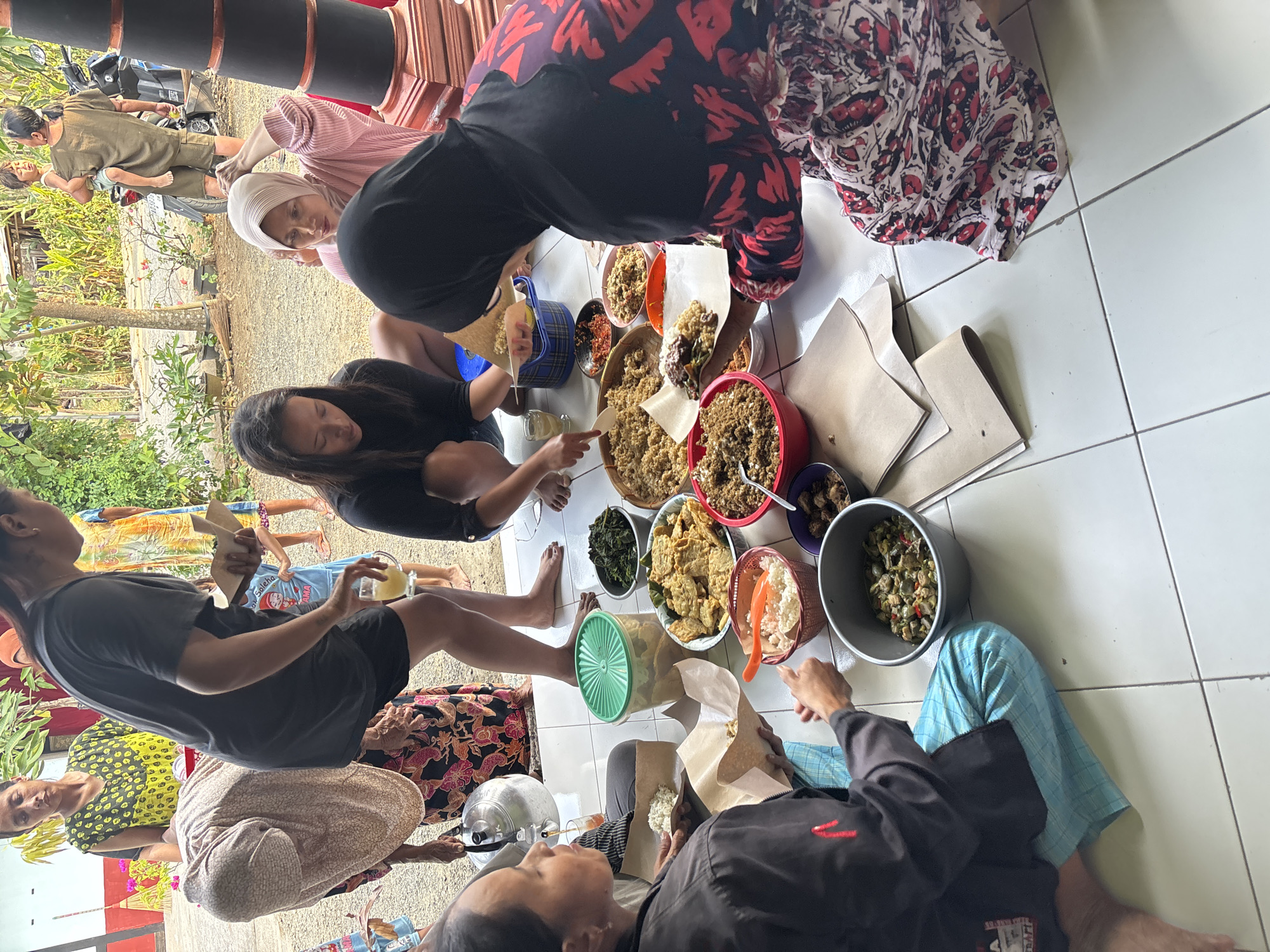 The image depicts an outdoor gathering in Tirtomoyo, Indonesia, where a group of people are seated on a tiled floor around a spread of food. Several individuals, mostly women, are visible, some in traditional attire and others in casual clothing. They are engaged in eating, serving, and interacting with each other. The food appears to be served family-style, with various dishes including rice, vegetables, and what looks like fried items, placed in bowls and platters on the ground. The setting suggests a residential or community area, with elements of Indonesian architecture and vegetation in the background. A motorcycle is parked nearby, indicating a rural or semi-urban environment. The overall atmosphere suggests a communal meal or celebration.