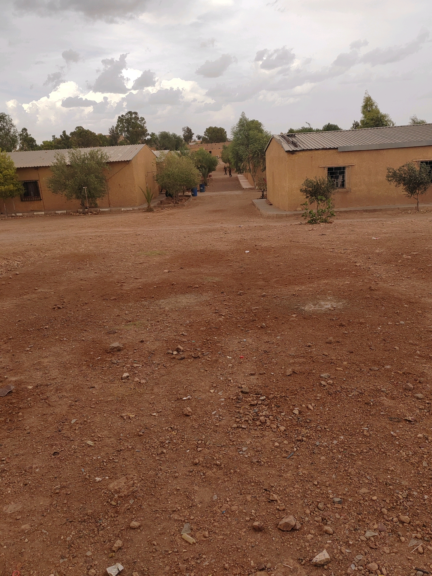 The image depicts an outdoor scene, likely in a rural or semi-urban area, characterized by a dusty, reddish-brown ground surface littered with small stones and some debris. In the background, there are several low-lying buildings with light-colored walls and corrugated metal roofs. Green trees and shrubbery are scattered around the buildings and along a pathway. The sky is overcast with dramatic, cloudy formations. The architecture of the buildings suggests a utilitarian purpose, possibly an educational or community facility. The overall impression is one of a dry, somewhat arid environment.