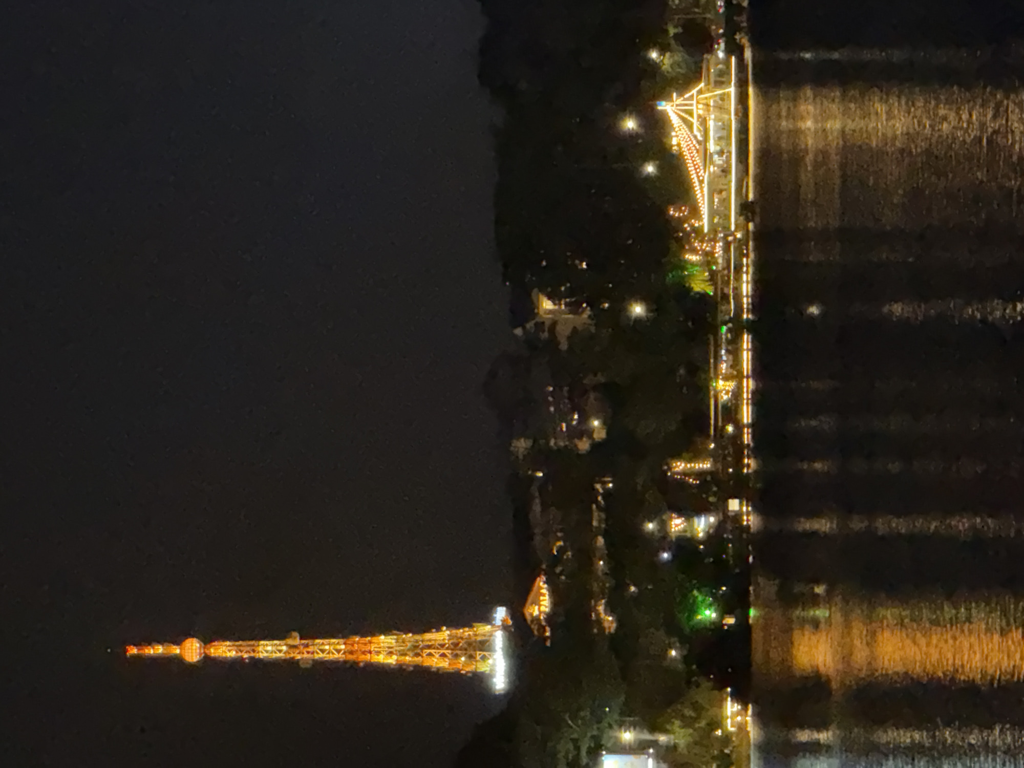 The image captures a nighttime scene along a waterfront in Da Lat, Vietnam. The most prominent object is a brightly lit tower, possibly a radio or observation tower, adorned with numerous orange and yellow lights that illuminate its lattice structure. The tower is reflected in the dark, rippling water, creating a shimmering effect.
Along the shoreline, a brightly lit structure resembling a promenade or a building with a festive atmosphere is visible. It is decorated with strings of white and orange lights, suggesting a celebration or a lively evening. The lights create a warm, inviting glow against the dark landscape and water.
Trees and foliage are interspersed along the shoreline, with some areas also featuring subtle lighting, hinting at a park or garden setting. The overall ambiance is one of enchantment and festivity, with the artificial lights contrasting sharply against the natural darkness of the night. The water appears calm, with reflections indicating a still night, possibly with a light breeze. The time of day is clearly night, as indicated by the dark sky and the illuminated man-made structures. There are no visible people in the frame, but the presence of the illuminated structures suggests human activity and a public gathering place. The scene evokes a sense of wonder and peacefulness. No text is visible in the image.