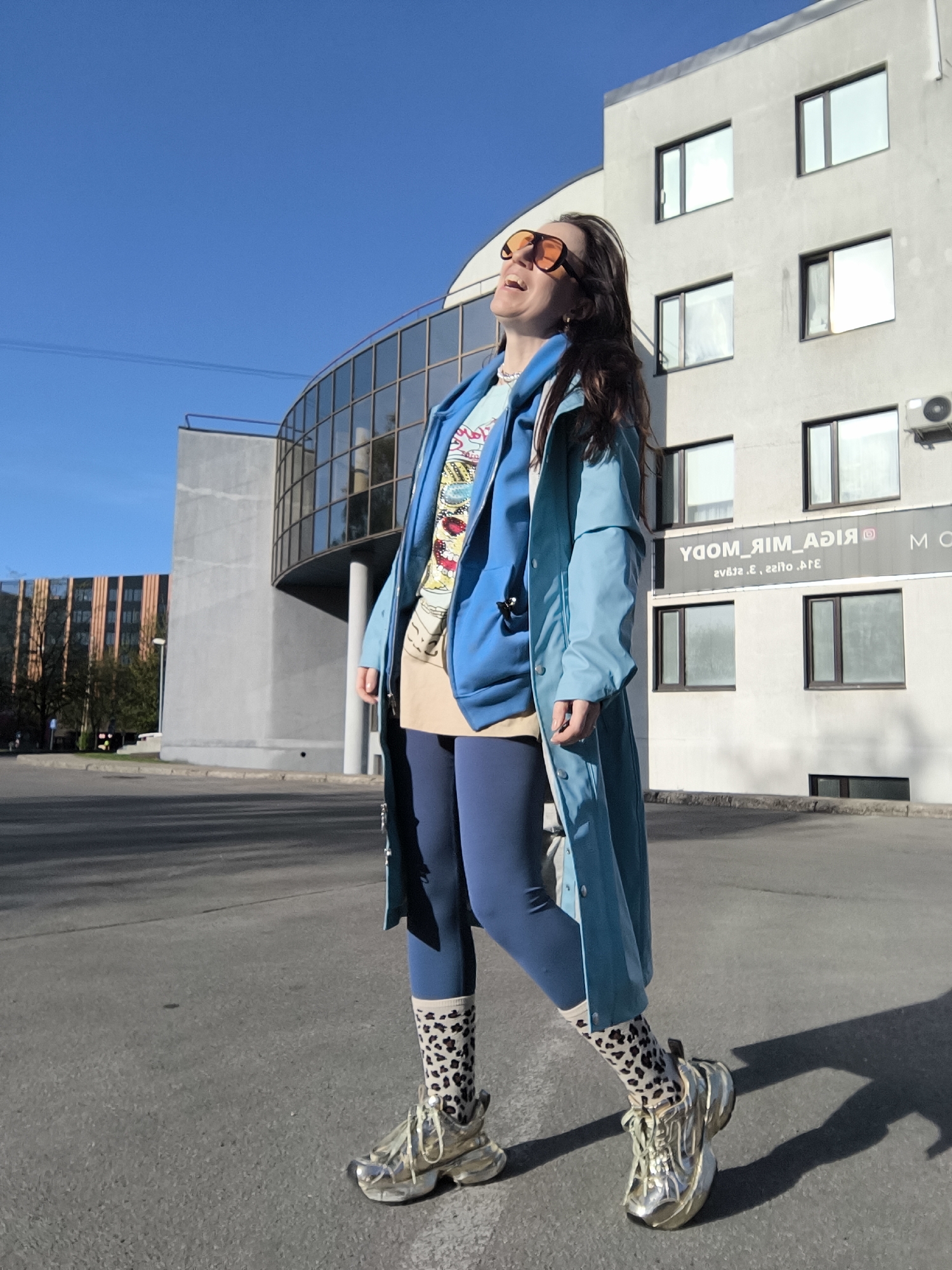 A medium shot depicts a woman standing outside against a backdrop of modern architecture in Riga, Latvia. The woman, with long brown hair and orange-tinted sunglasses, is laughing with her head tilted upwards. She is wearing a layered outfit that includes a light blue trench coat, a blue hoodie, a patterned shirt, a beige skirt, and blue leggings. Her footwear consists of leopard-print socks and shiny gold sneakers. The buildings behind her include a curved, glass-fronted structure and a white building with windows, bearing the reversed text "RIGA MIR MODA" along with other text, presumably a business name or advertising. The area appears to be a paved parking or street area under a clear blue sky.