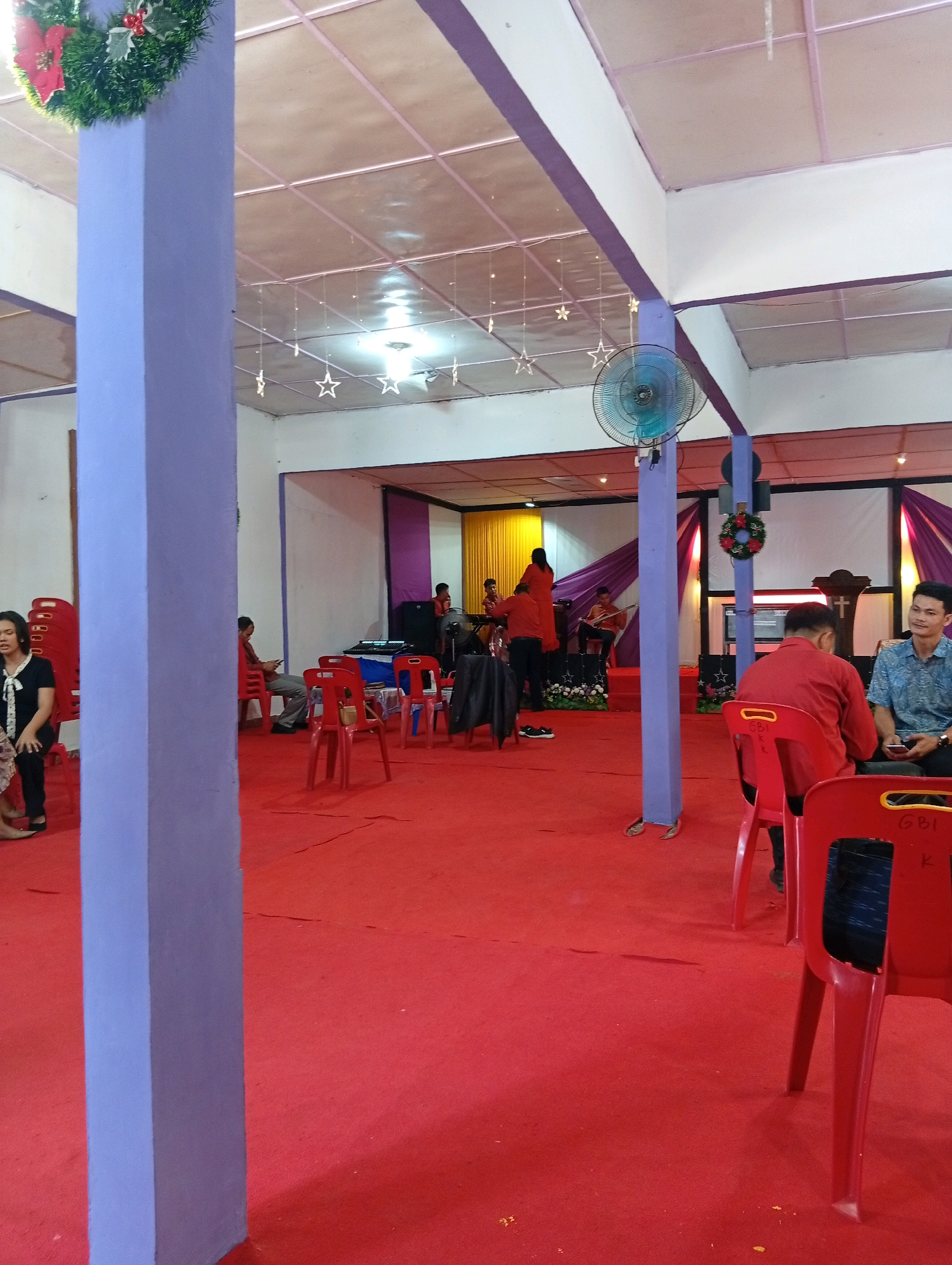 The image depicts an indoor gathering at what appears to be a church or community hall in the City of Gunungsitoli, Indonesia. The space is carpeted in vibrant red, with purple and white walls and ceiling. A large purple pillar stands prominently in the foreground. Several red plastic chairs are arranged throughout the room.
In the background, a small stage area is set up with purple and yellow drapes. Some individuals are seated on the stage, and at least two are playing musical instruments, possibly guitars. A keyboard is also visible on the stage.
On the right side of the image, two men are seated in red chairs facing each other, engaged in conversation. One man is wearing a blue patterned shirt, and the other a red shirt. In the left foreground, a woman in a black top and patterned skirt is seated, looking towards the left.
Decorative elements include a Christmas wreath hanging from a pillar and small star-shaped lights suspended from the ceiling. A ceiling fan is also visible. The overall atmosphere suggests a religious or community event taking place.
