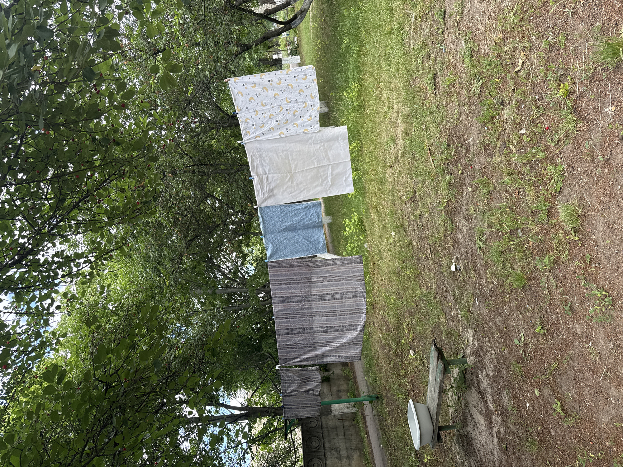 The image shows laundry hanging on a clothesline in an outdoor setting. The clothesline is strung between two poles and stretches across a grassy area with trees. Several pieces of laundry are visible, including towels and sheets, with varying patterns and colors. In the foreground, there is a small wooden bench with a white basin on top of it. A concrete wall runs along the left side of the image, behind the clothesline. The environment appears to be a residential area with lush green trees and vegetation.