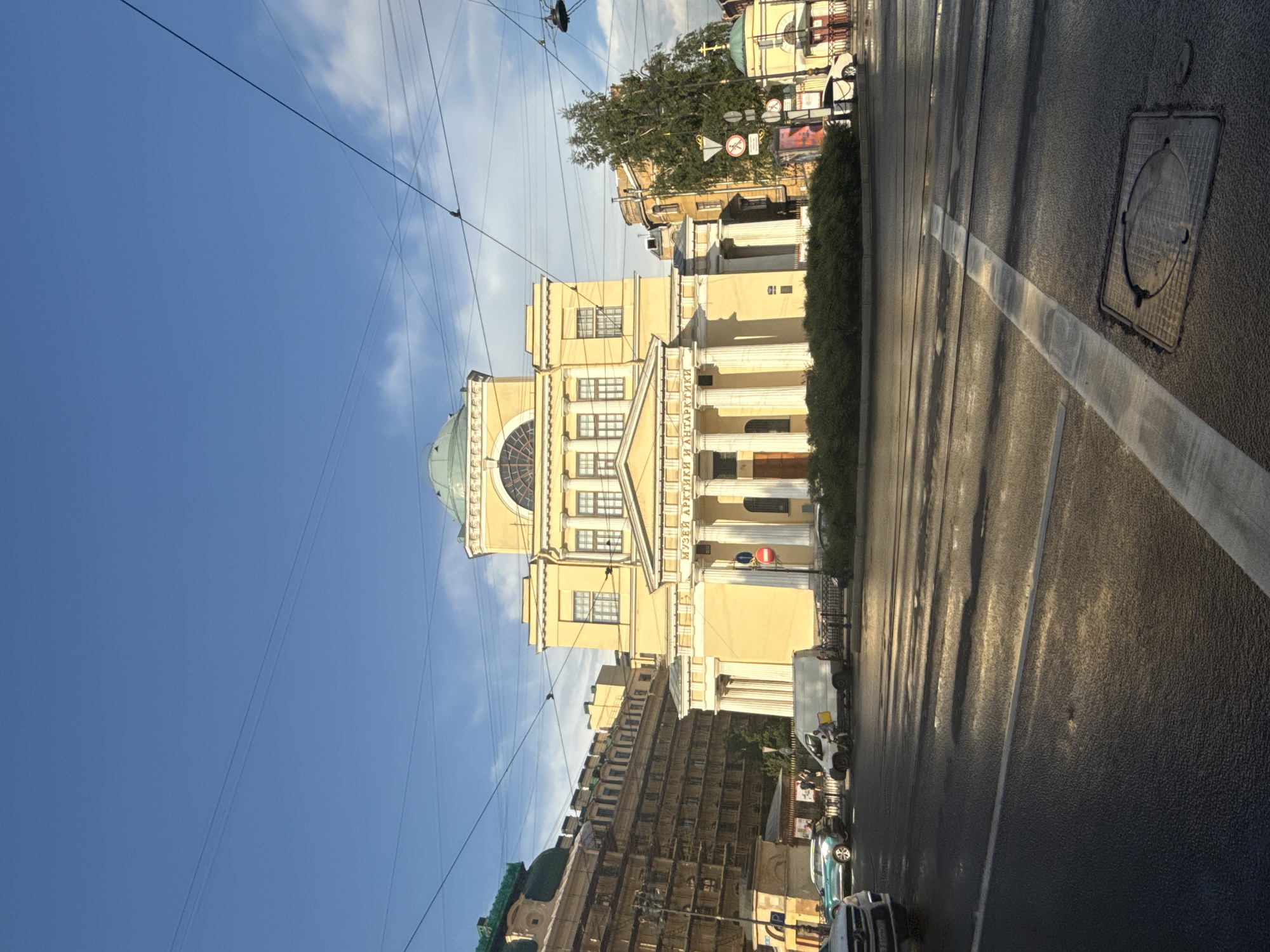 The image shows a street scene in Saint Petersburg, Russia, featuring the front facade of a yellow neoclassical building with a large dome. The building has a prominent portico with white columns and a pediment. Above the portico, there is an arched window and several rectangular windows on the upper floors. The text "МУЗЕЙ АРКТИКИ И АНТАРКТИКИ" (Museum of Arctic and Antarctic) is visible on the building's facade.
In the foreground, a wet asphalt street is visible with white lines marking lanes and a pedestrian crossing. Several vehicles are present on the street, including a white van, a blue car, and a black car. A cyclist is also visible near the white van. Overhead, power lines and trolley wires crisscross the sky.
To the left of the main building, another large building with a green roof is partially visible. To the right, a smaller yellow building with a green dome and a cross on top is visible, suggesting it might be a chapel or part of a religious complex. Green bushes line the front of the main building. The sky is blue with scattered clouds. The overall impression is of a bright, sunny day, with the street appearing wet, possibly from recent rain.