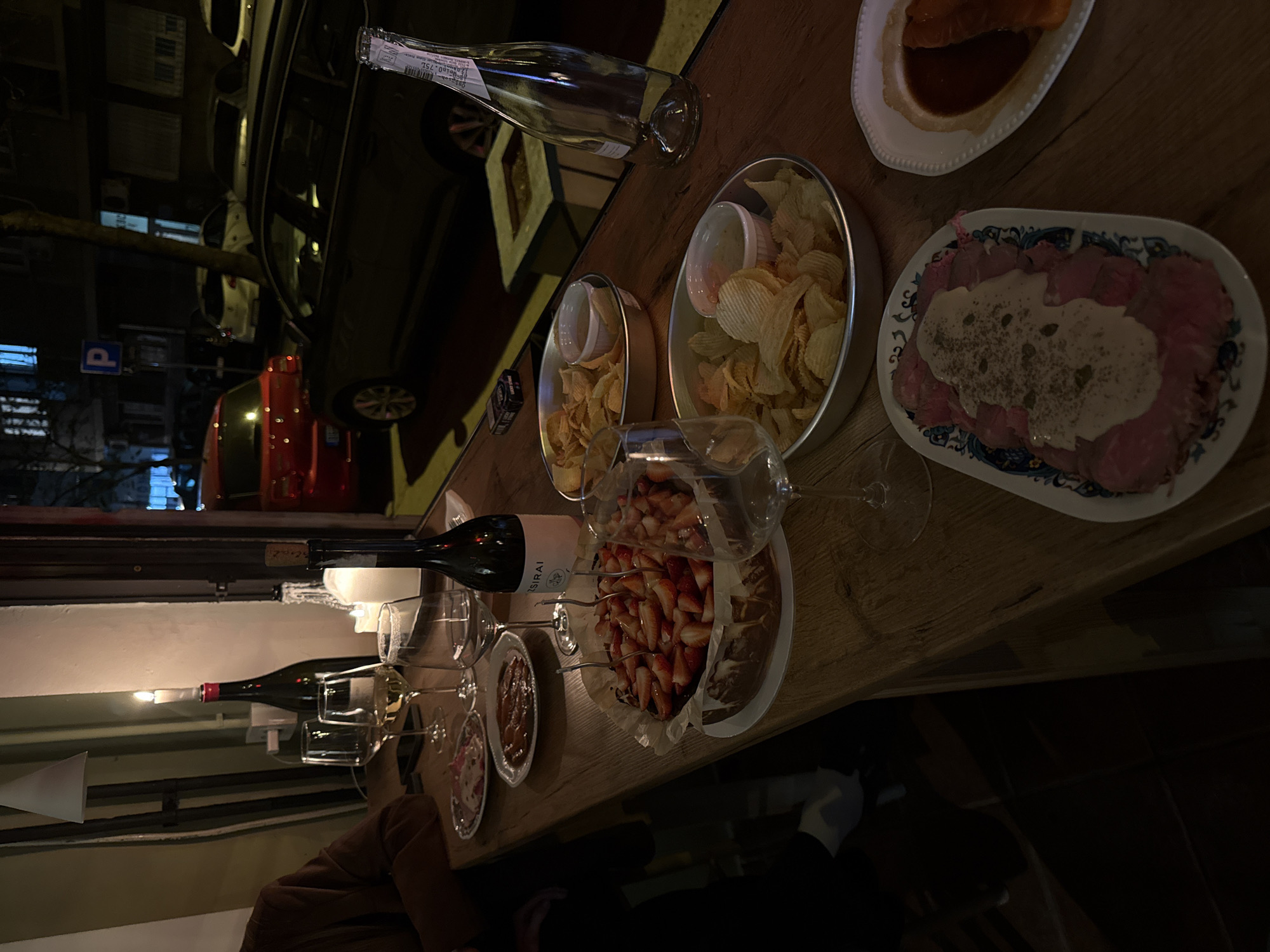 The image depicts a table laden with food and drink in what appears to be a restaurant or bar in Belgrade, Serbia. The focus is on a selection of appetizers, including potato chips, a carpaccio dish, and another dish with a dark sauce. A strawberry tart sits closer to the foreground, alongside a bottle of wine labeled "Sirai" and several wine glasses. Another bottle is present but its label is illegible. Through a nearby window, one can see streetlights, parked cars, and an indistinct red vehicle, suggesting the establishment is street-facing. A man's shoulder can be seen in the lower left-hand corner of the shot. The environment is dimly lit.
