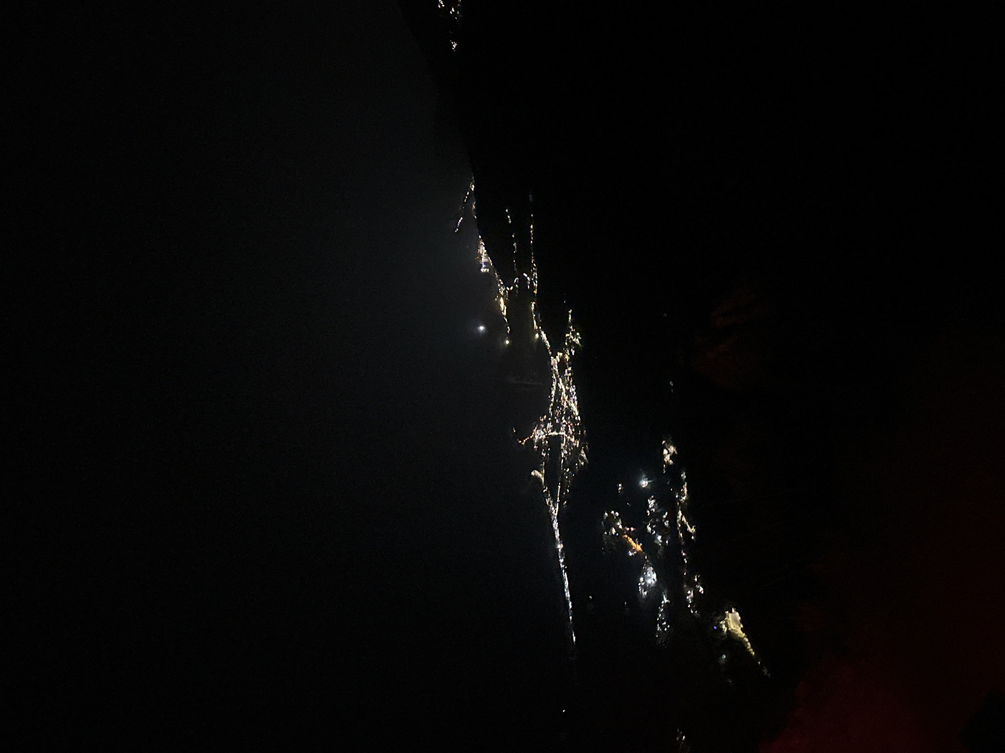 The image shows an aerial view of the city of Gluhi Do, Montenegro at night. The city lights are visible in the darkness, spread out across the landscape. The lights appear to be concentrated along a coastal area and extending inland. A bright moon is visible in the upper left quadrant of the image. The foreground is dominated by darkness, with hints of dimly lit structures or terrain. The overall scene depicts an urban area illuminated at night from a high vantage point.