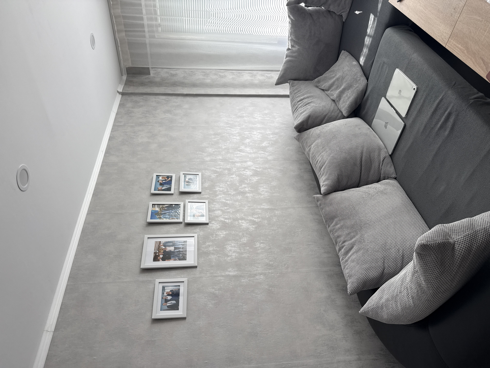 The photograph shows an interior space with grey tiled flooring and a light grey wall to the left, contrasted by a darker grey sofa positioned on the right side of the frame. Natural light enters the room through a window covered with white curtains, casting shadows on the floor near the window. The sofa is adorned with multiple square pillows in a matching grey fabric and two white tablets are placed on it. On the wall, a series of five framed photographs are arranged vertically, each containing images of people. The room features a recessed lighting fixture in the wall. The photograph was taken in Lovech, Bulgaria.