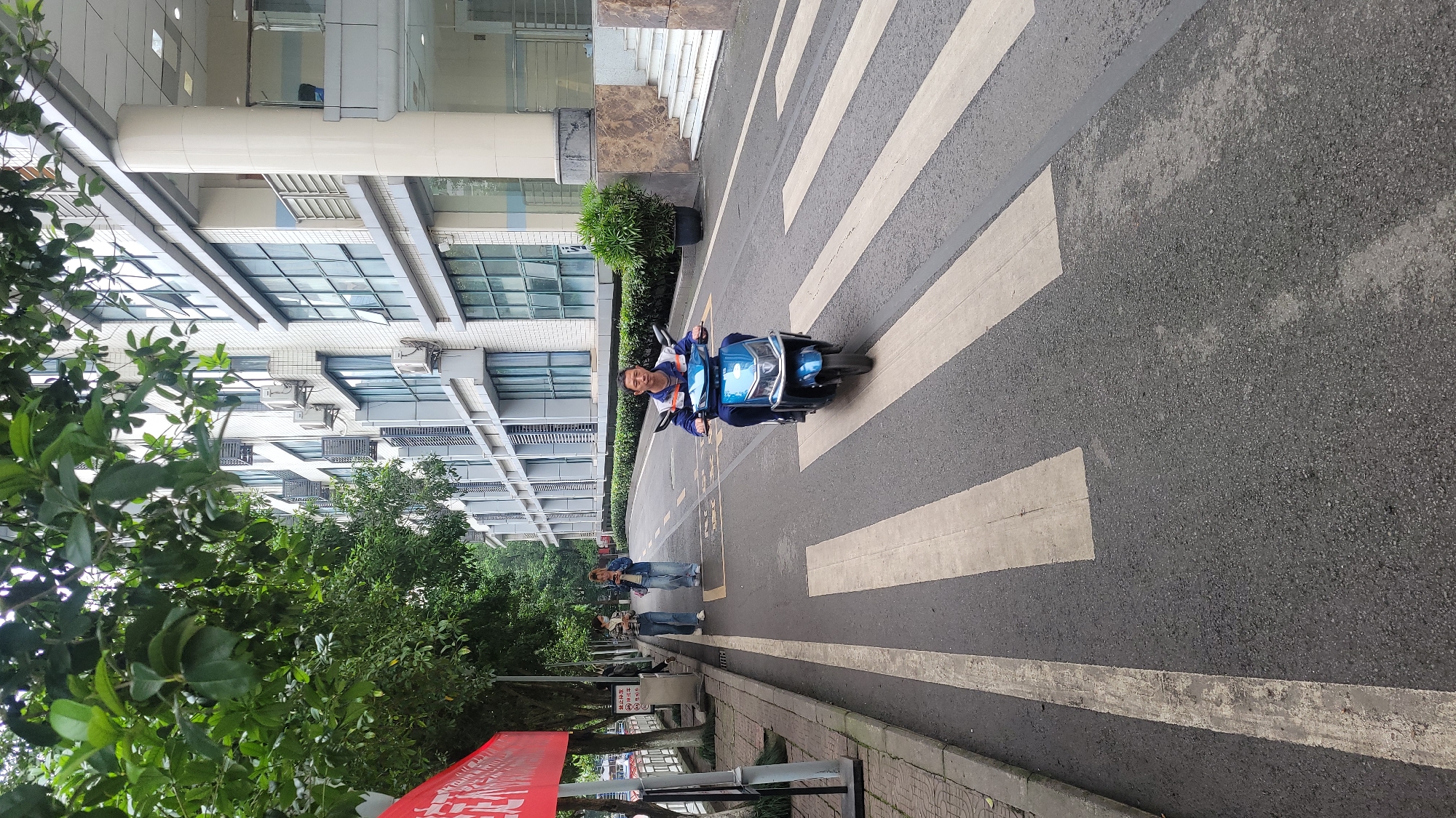 The image captures a street scene in Yuzhong District, China, with a man riding a blue electric scooter across a crosswalk. The crosswalk is marked with white stripes on an asphalt road. In the background, modern buildings with glass windows and brick facades are visible, adorned with air conditioning units. Trees and bushes line the street, adding greenery to the urban environment. Two people can be seen walking on the sidewalk in the distance. A red banner is partially visible on the left side of the image, next to a signpost with Chinese characters. The lighting suggests it is daytime.