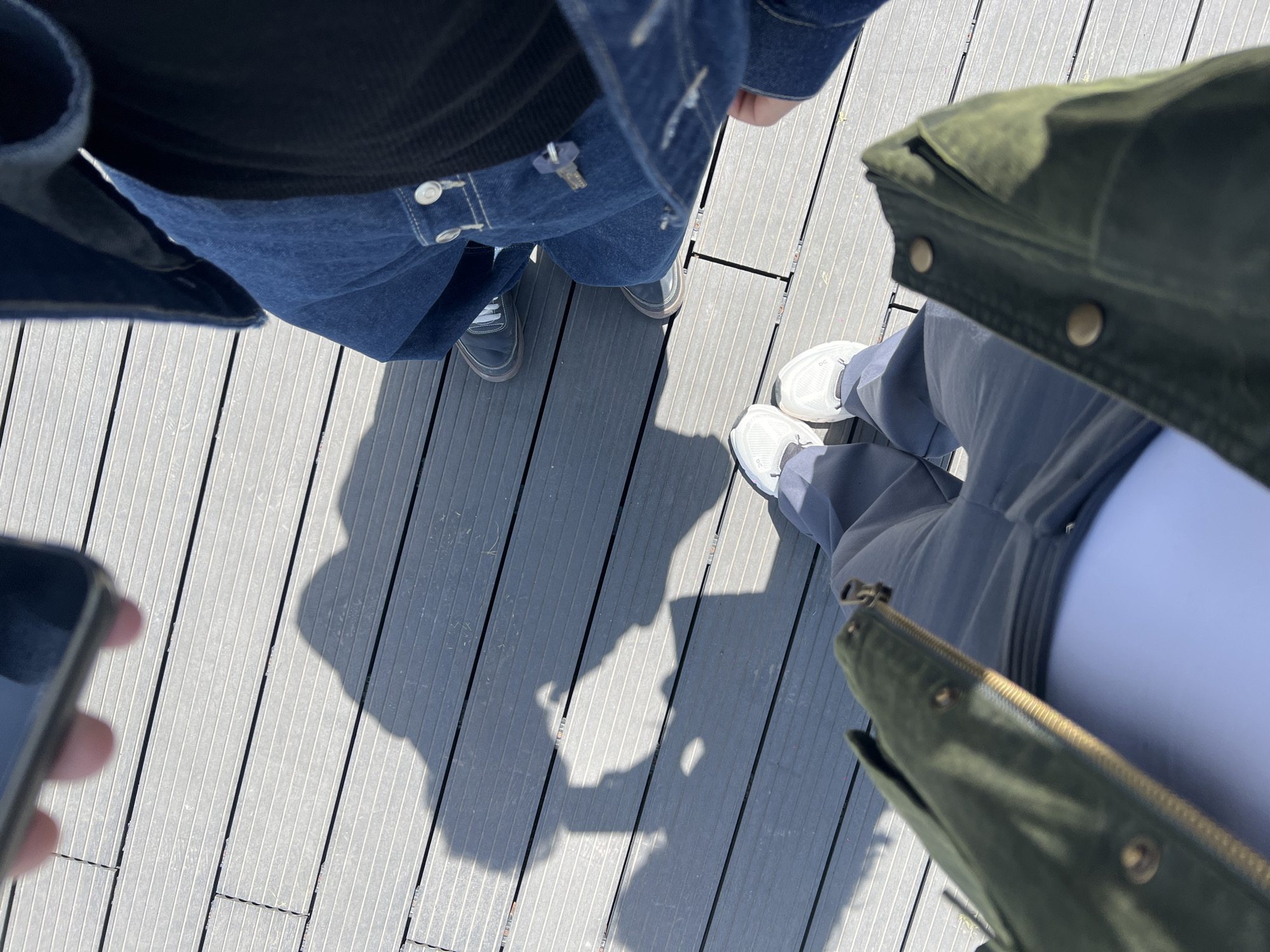 The image shows a top-down view of two people standing on a gray, wooden-slatted surface. The person on the left is wearing a blue denim jacket over a black shirt, and blue jeans, with dark-colored shoes. The person on the right is wearing an olive green jacket and light blue pants with white sneakers. A smartphone is visible at the bottom left corner. The scene is brightly lit, casting distinct shadows of the individuals onto the wooden surface. The location is Unknown, Unknown.