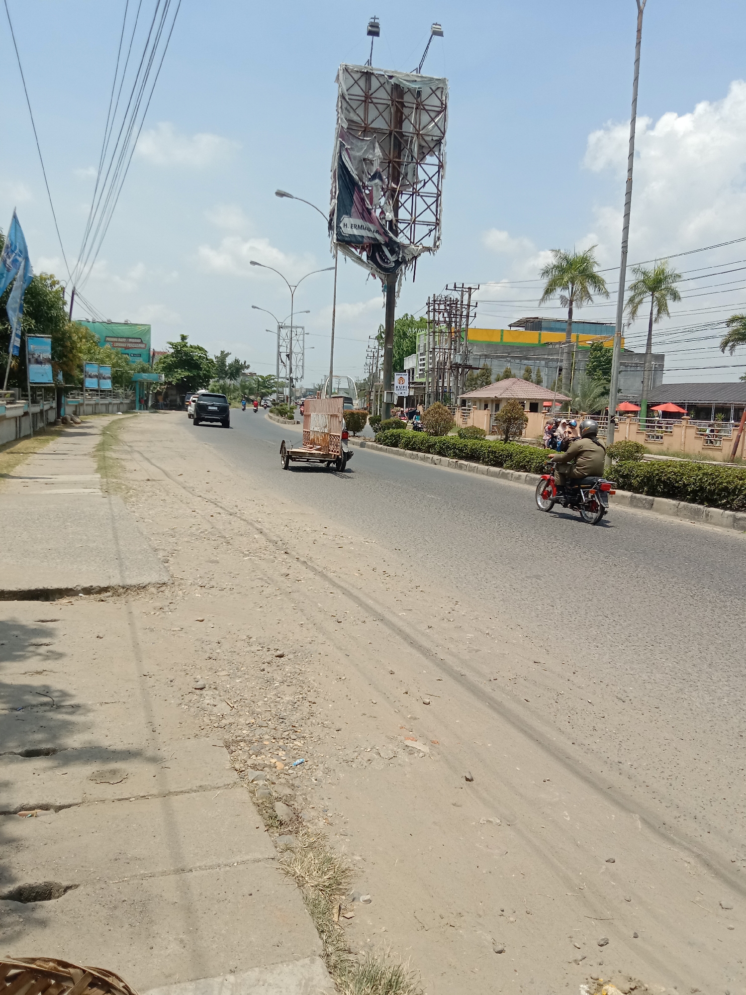 The image captures a sunlit street scene in Lhokseumawe, Indonesia, on a clear day with a bright blue sky dotted with scattered white clouds. The main thoroughfare is paved, supporting vehicular traffic, while the immediate left side features a rough, unpaved dirt shoulder and a broken concrete sidewalk.
In the mid-ground, traffic moves along the road. A person wearing a helmet and a green jacket rides a red motorcycle away from the viewer. Ahead of them, a three-wheeled motorized vehicle, likely a motorcycle adapted with a sidecar or trailer, carries what appears to be wooden crates or a similar cargo. Further down the road, a black SUV is also visible among other vehicles.
The scene is framed by various structures and elements. A large, dilapidated billboard with a rusty metal frame and torn advertising material stands prominently on the right side of the road. Visible text on the billboard reads "H. ERMIAWAN ARE". Numerous overhead power lines crisscross the sky, connected to utility poles that line the street. Streetlights are also interspersed. Along the roadside, there are buildings, including one with a distinctive yellow and green upper section and another with a reddish-brown tiled roof further back. Lush green bushes, palm trees, and other foliage are scattered throughout the environment, providing some greenery amidst the urban setting. The unpaved areas are dusty and uneven, with small rocks and patches of dry grass. On a green sign further down the street, obscured text partially reads "PUDING BARU / MINUMAN."