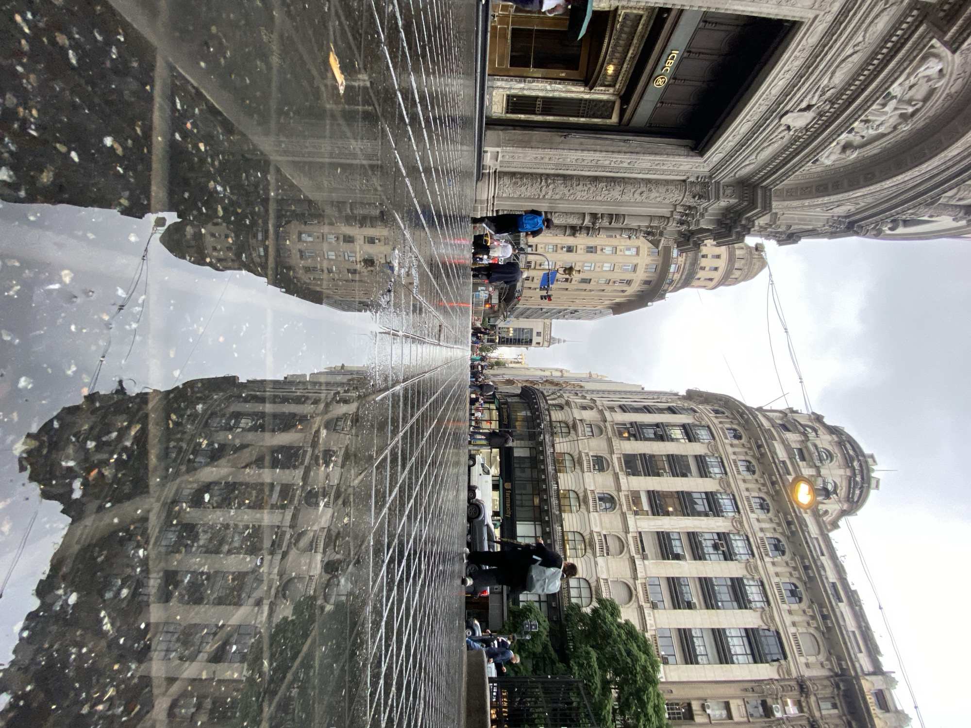 The image depicts a street scene in Buenos Aires, Argentina, likely near Avenida Corrientes. The perspective is from a low angle, showing a reflection in a wet or polished surface, possibly wet pavement or a reflective plaza. The reflection mirrors the architecture and sky. Buildings with classical facades and arched windows line both sides of the street, one with the ICBC bank logo visible above an entrance. People walk along the street and are captured both in reality and reflection, creating a layered visual effect. Cars are visible parked along the street. The sky appears overcast.