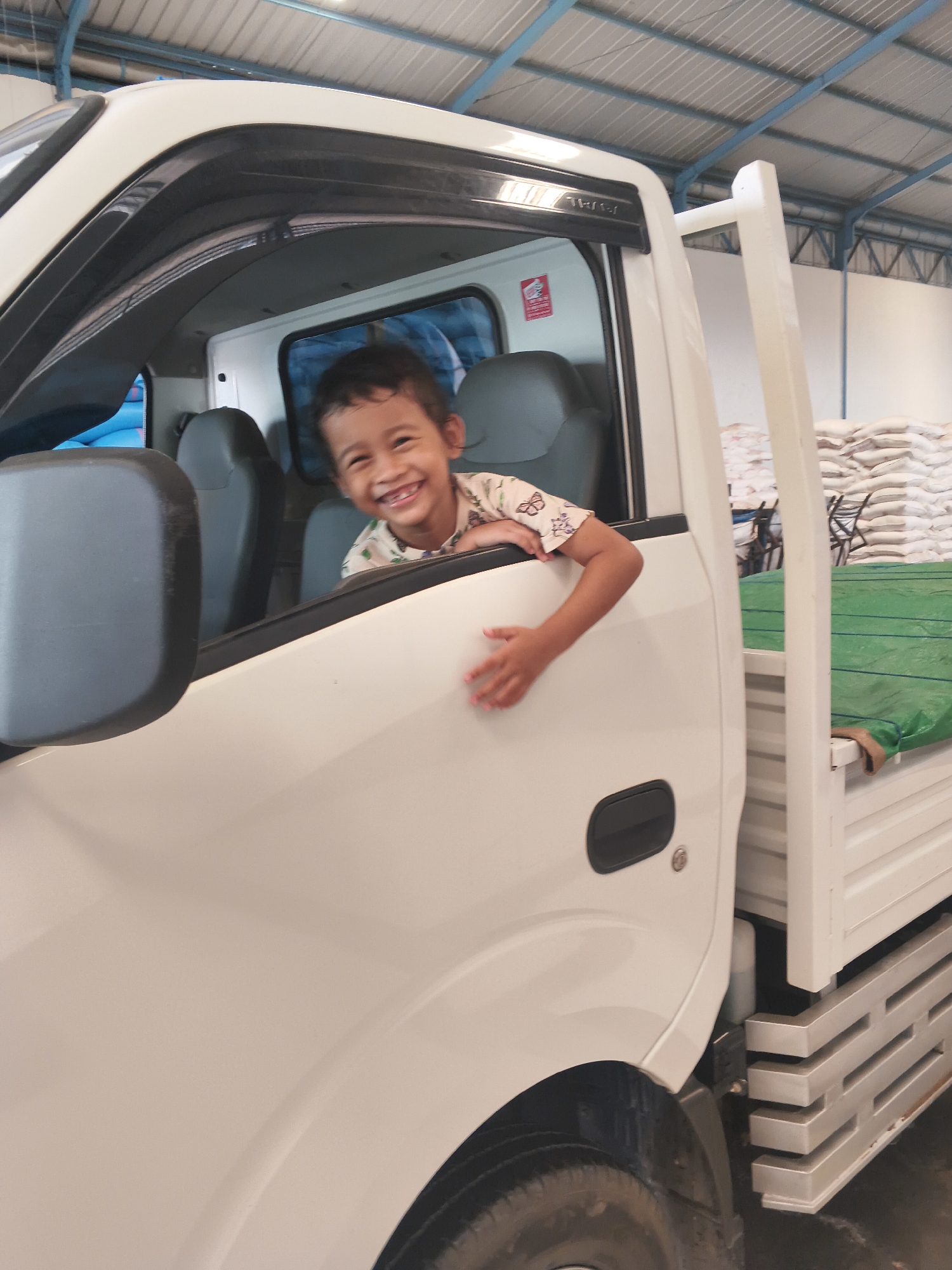 A young boy with dark hair smiles broadly as he leans out of the open window of a white truck. He is wearing a light-colored shirt with a pattern of small butterflies. The truck's interior, including a gray seat, is visible behind him. The truck is parked inside a large, open building with a corrugated metal roof supported by blue and white trusses. Stacks of white bags are visible in the background, along with a green tarp covering a portion of the truck bed. The image appears to have been taken in Bandar Lampung, Indonesia.
