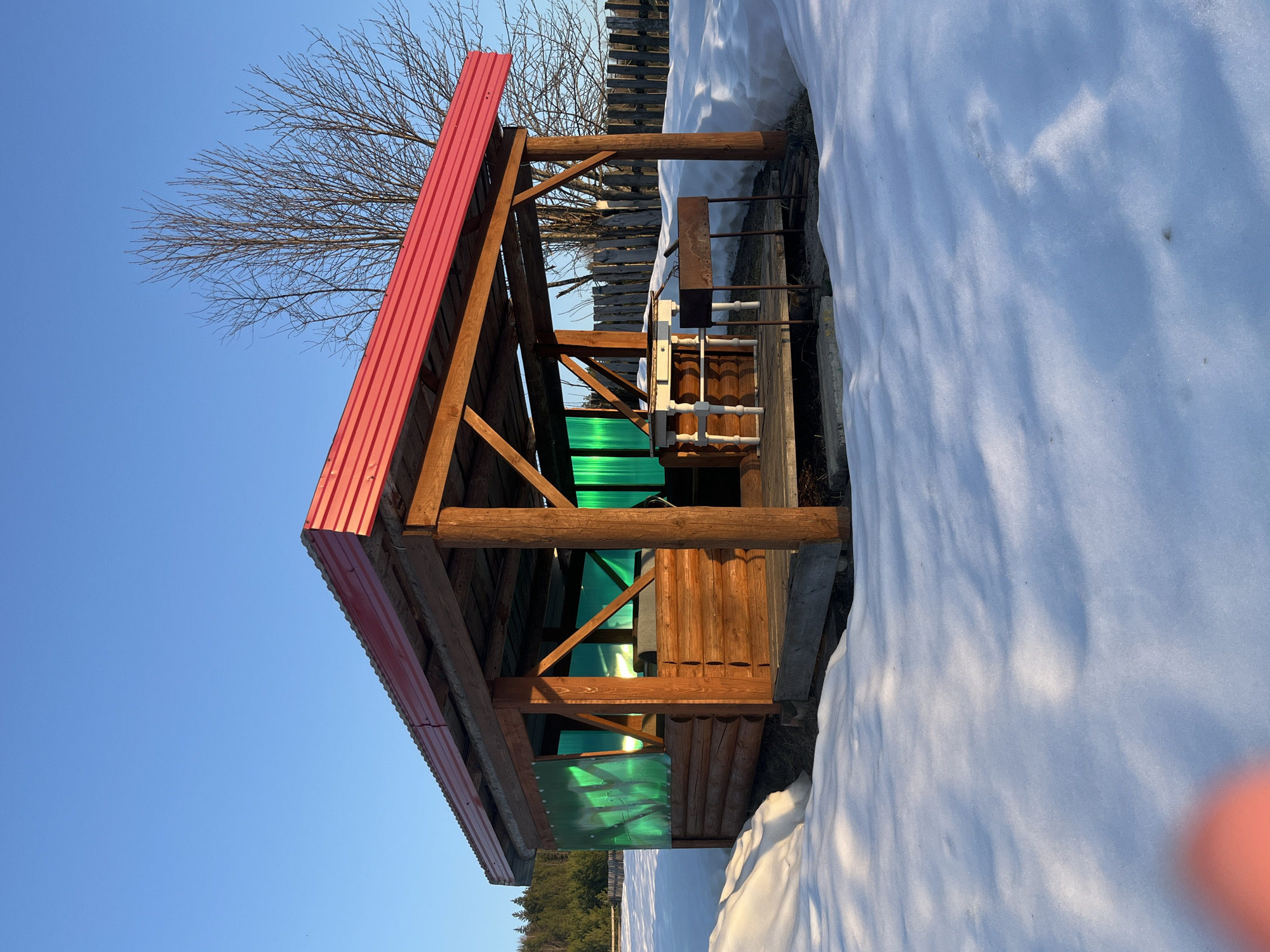 The image shows a wooden structure with a red metal roof in Большой Букор, Russia. The structure has green translucent panels on the sides. There's snow on the ground next to the structure, and a bare tree is visible in the background. The sky is clear and blue. Part of a wooden fence can be seen behind the structure. There is a device with pipes next to the structure on the snow side.
