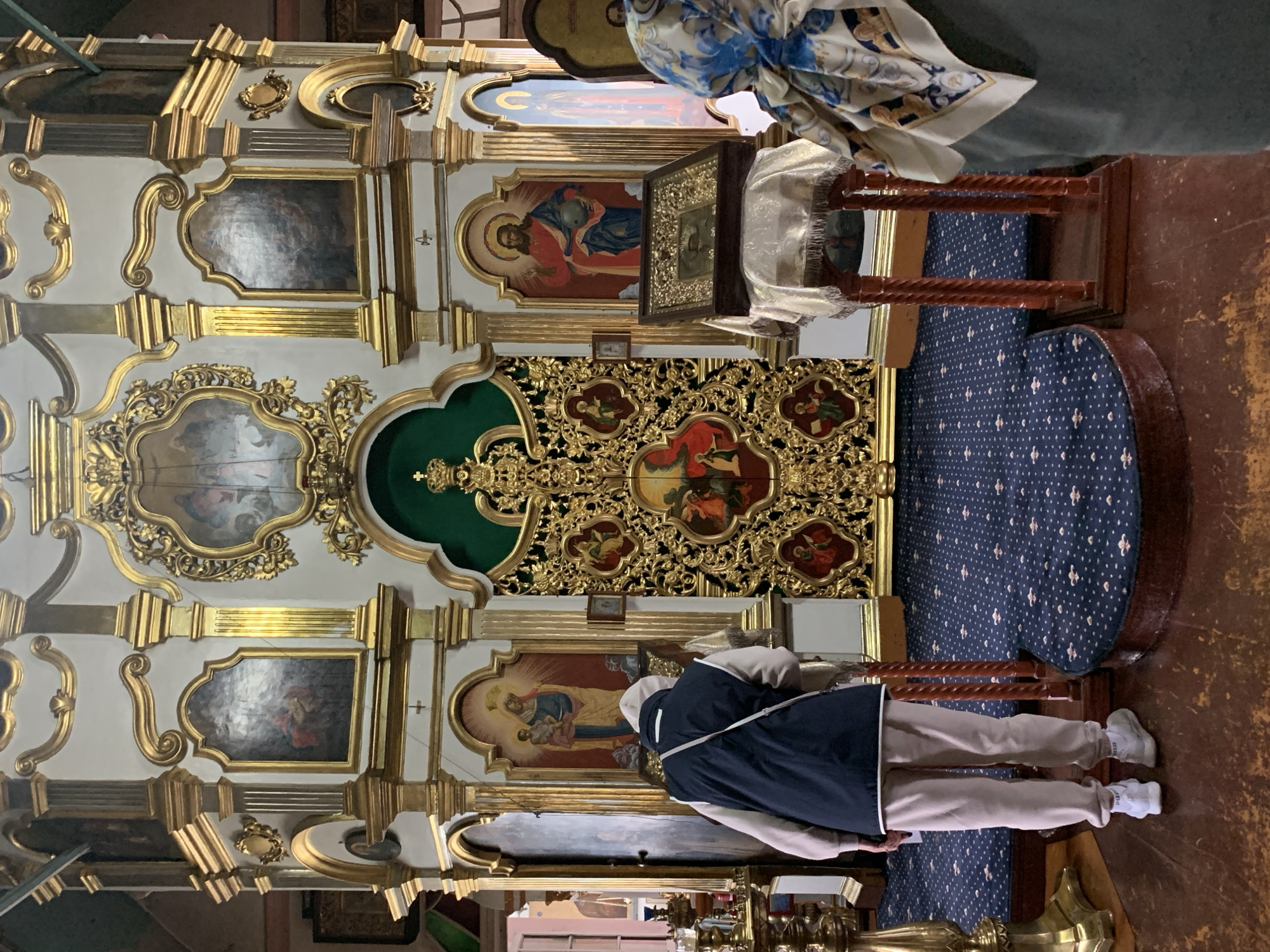 The image shows the interior of an Orthodox church, likely the Cathedral of the Assumption of the Virgin Mary in Oryol, Russia, given its opulent gilded iconostasis and traditional iconography. Two individuals are present. In the foreground, a person from the back is wearing a light-colored hooded sweatshirt, a dark vest, and light-colored pants, facing a large, ornate golden iconostasis. They appear to be looking at or praying before an icon displayed on a stand in front of the iconostasis. To the right, another person, mostly out of frame, is seen from the side, wearing a patterned headscarf. The iconostasis itself is a multi-tiered structure adorned with numerous gold-leaf decorations and religious paintings, typical of Eastern Orthodox churches. The floor is covered with a dark blue carpet featuring a repeating white pattern. The lighting suggests an indoor environment, possibly with natural light filtering in from unseen sources.