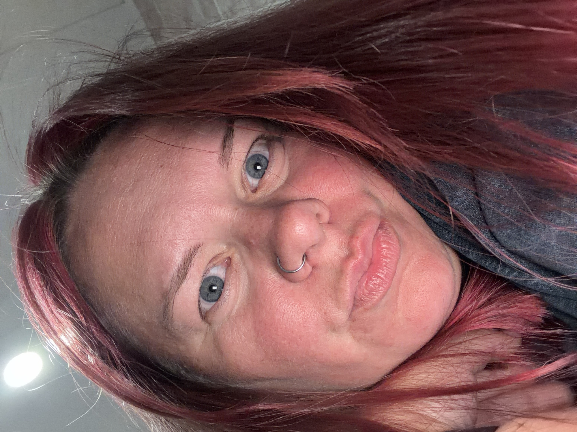 A close-up, tilted self-portrait of a woman with red-toned hair, blue eyes, fair skin, and a silver nose ring. She is wearing a denim shirt. The location in the city of Chaikovsky, Russia is not discernible from the image.