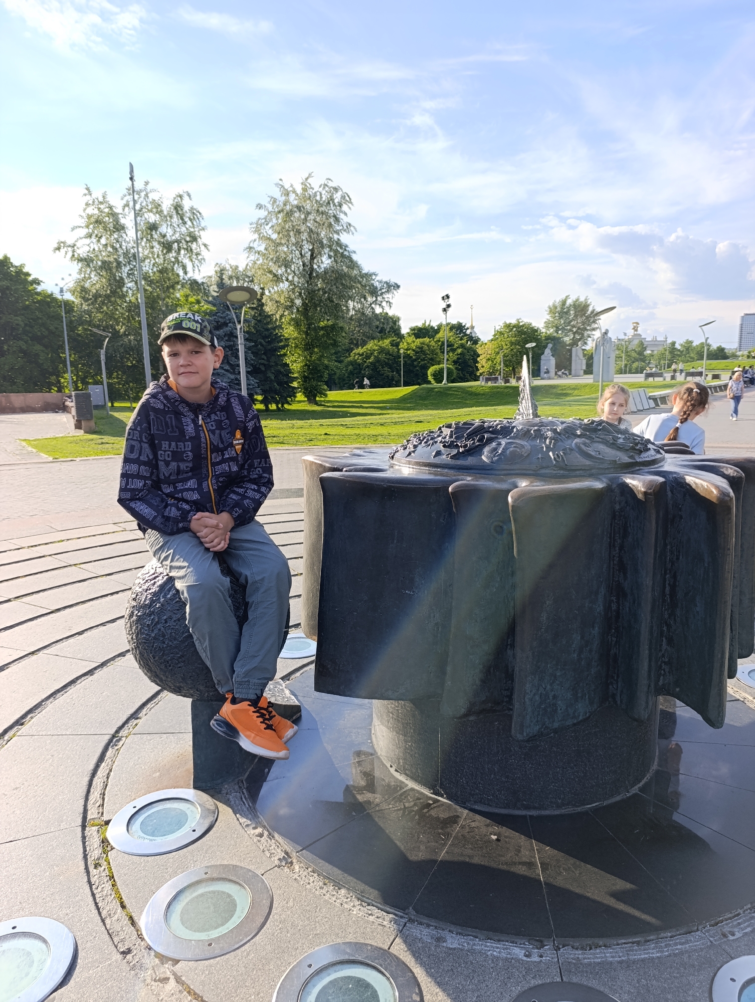 A bright outdoor scene captures a young boy and two girls interacting with a large, elaborate sundial in what appears to be a public park or square in Moscow, Russia.
In the foreground, a young boy with a neutral expression is seated on a dark, textured spherical object. He wears a camouflaged baseball cap with "FREAK 001" visible on the front, a dark patterned jacket with fragmented English words and phrases like "HARD GO" and "CANNOT", grey pants, and striking orange and white sneakers. His hands are clasped in front of him.
To his right, a monumental, dark metallic sculpture dominates the midground. It features a circular top intricately decorated with raised patterns, likely representing celestial motifs or a calendar, and a central gnomon typical of a sundial. The base of the sculpture is fluted or ribbed. Sunlight catches its surface, creating a sunflare effect. Around the base of this sculpture and the spherical seat, the paved ground has concentric circular patterns embedded with circular lights.
Behind the sundial, two young girls are partially visible, looking towards the right. One has blonde hair, the other brown hair tied in pigtails. They appear to be observing their surroundings.
The background reveals a lush green park landscape with various trees, including tall deciduous and coniferous types. Lampposts and park benches are scattered throughout. In the far distance, blurred structures suggest an urban setting, including a building with a golden spire, possibly indicating a landmark within Moscow. The sky is bright with scattered white clouds, and the sun, coming from the upper left, casts distinct shadows. The overall atmosphere is leisurely and bright.