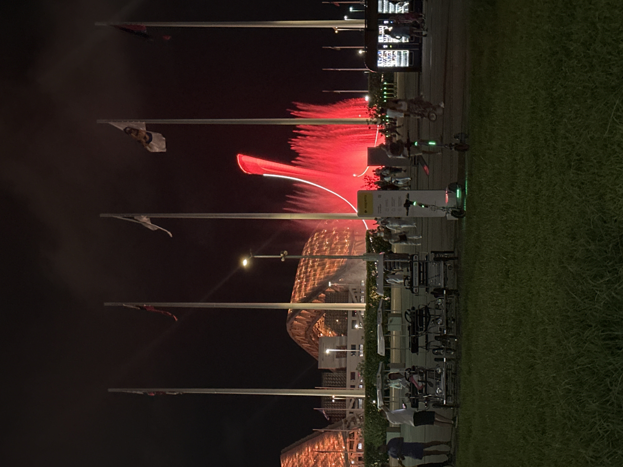 At night, a vibrant red fountain sprays water upwards in front of a large, modern building with an arched, orange-lit roof. Several flagpoles with flags are visible against the dark sky. In the foreground, there are several multi-person pedal carts and two electric scooters. People are walking around, some are near the pedal carts, others are near a vending machine. The area is lit by artificial lights, including streetlights and the illuminated building and fountain. The ground is paved in some areas and covered with grass in others.