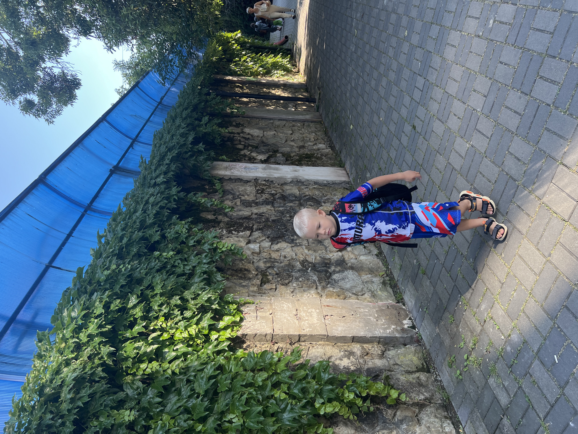 In Sochi, Russia, a young boy with blond hair walks along a paved pathway bordered by a stone wall covered in ivy. He wears a blue and red patterned shirt and shorts, along with black sandals and a backpack. Above the wall, a blue corrugated roof structure extends over the pathway. In the background, two women are visible walking further down the path, and trees line the right side of the scene. The sun casts shadows on the pathway, indicating a daytime setting.