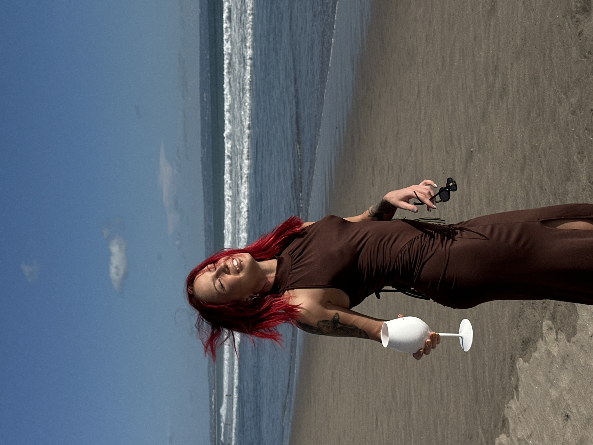 A woman with vibrant red hair and tattoos is standing on a sandy beach in North Kuta, Indonesia. She is wearing a brown dress and holding a white wine glass in one hand and a pair of sunglasses in the other. The ocean is to her left, with waves visible in the background. The sky is blue with some clouds. The scene suggests a relaxed, carefree atmosphere.