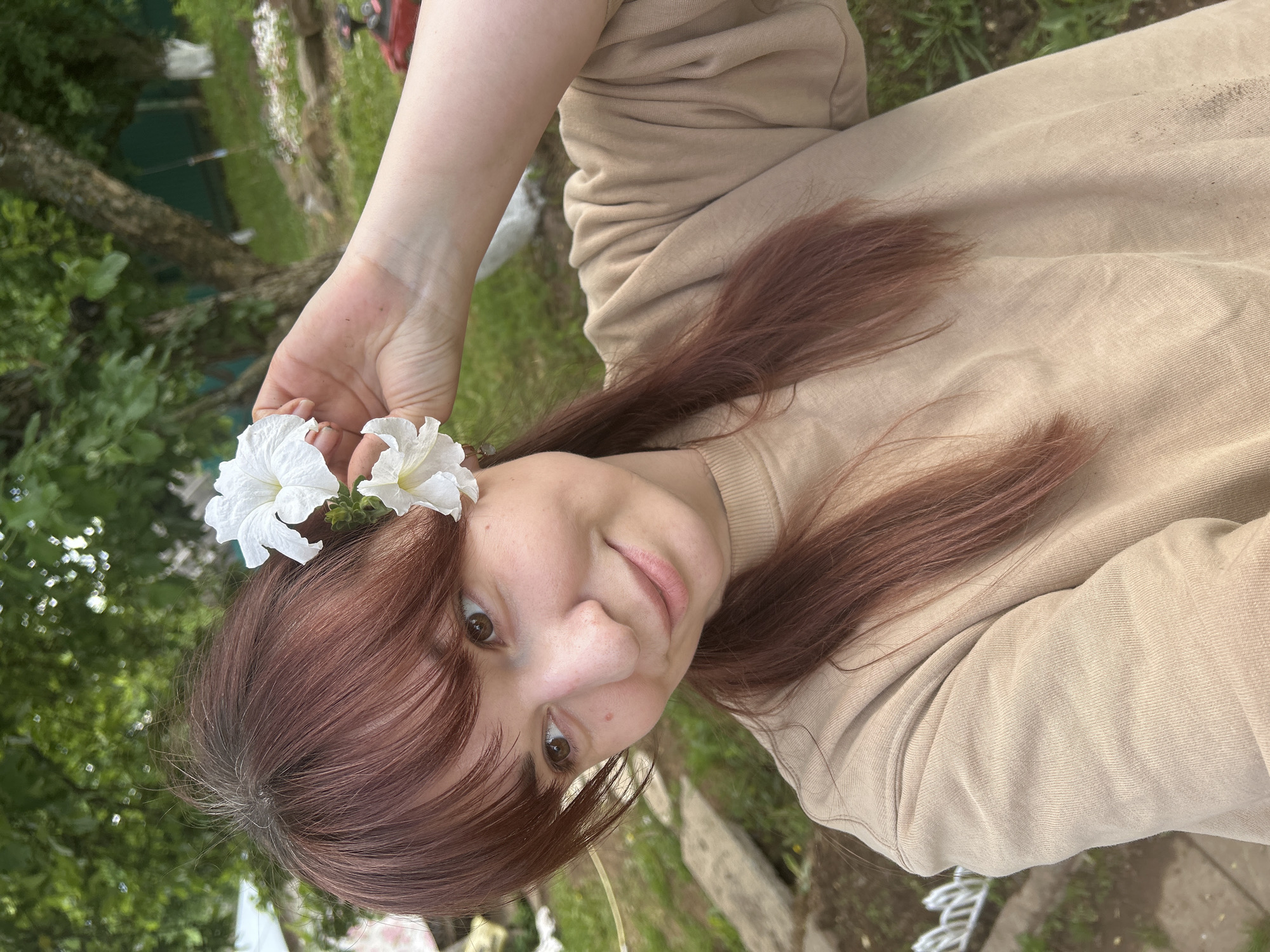 A woman with brown hair and brown eyes is holding two white flowers near her forehead. She is wearing a beige sweatshirt. The background appears to be outdoors, with greenery and possibly a lawn. The location is Фоки, Russia.