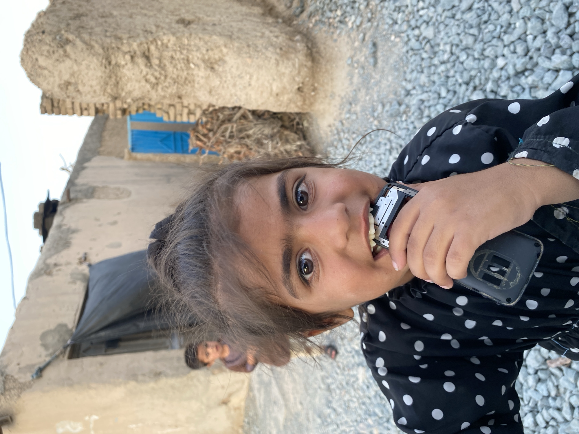 This media file depicts a young girl in an outdoor setting in Kandahar, Afghanistan. She is the primary subject, shown from the chest up, with her face turned towards the camera and tilted to her left. The girl has dark hair, prominent eyebrows, and brown eyes. She is wearing a black dress with white polka dots. In her right hand, she is holding a black object, possibly a mobile phone, with a silver-colored component near her mouth. Her mouth is slightly open, and she appears to be biting or touching the object with her teeth.
The background suggests a rural or semi-rural environment. To the left of the girl, a beige, textured building wall is visible. Further in the background, a blue door or window frame stands out against a wall. The ground is covered with gravel and small stones. In the upper left portion of the image, a rough, earthy structure, possibly a wall or roof, is prominent. Another child is partially visible in the background, to the lower left of the main subject, wearing a purple top.
The lighting suggests it is daytime, likely late afternoon or early evening, given the angle of the light and the shadows. The overall atmosphere appears casual and candid. The girl's expression is difficult to fully ascertain due to the way she is holding the object, but there's an element of curiosity or playfulness in her gaze and posture. The setting appears to be a residential area with simple construction typical of the region. No visible text is present in the media file.