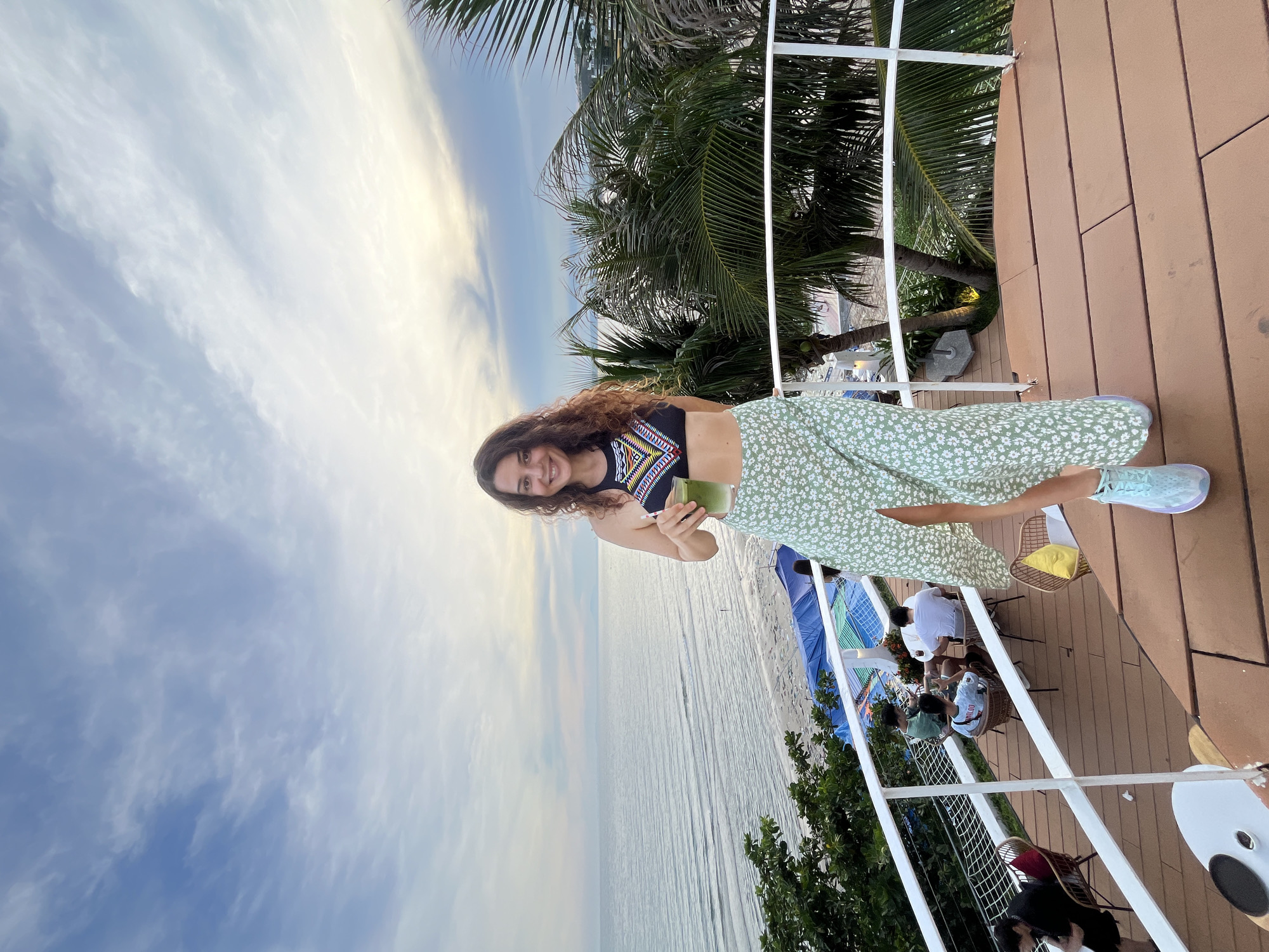 In Mũi Né, Vietnam, a woman stands on a wooden deck, holding a green drink, with the ocean and sky in the background. She wears a black patterned bikini top and a light green floral skirt with a slit, and light blue sneakers. In the middle ground, palm trees line a sandy beach with a blue tarp and people seated at tables. The sky is cloudy with a hint of sunset.
